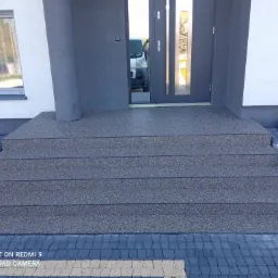 Schody wejściowe do domu wykończone posadzką żywiczną w kolorze szarym, dopasowane do elewacji i kostki brukowej.