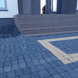 Ułożona kostka brukowa w odcieniach szarości i beżu przed wejściem do budynku z trzema schodami pokrytymi kruszywem.