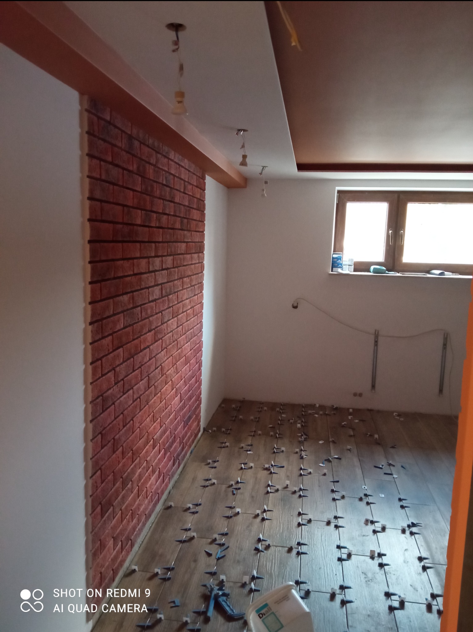 Montaż płytek imitujących cegłę na ścianie oraz układanie drewnopodobnych płytek podłogowych z użyciem krzyżyków dystansowych w trakcie remontu pokoju.