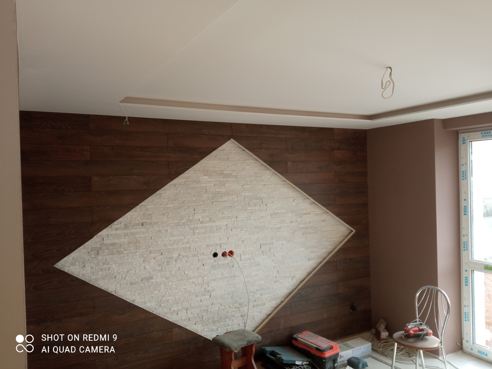 Ściana wykończona drewnopodobnymi panelami z wkomponowanym trójkątnym elementem z jasnego kamienia dekoracyjnego. Widoczne gniazdka elektryczne, narzędzia i krzesło w trakcie prac wykończeniowych.