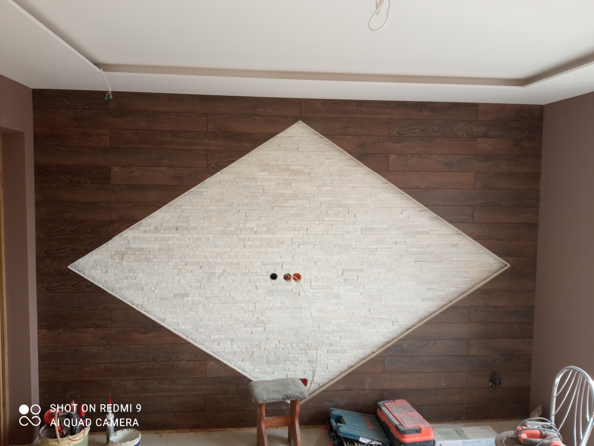 Ściana z dekoracyjnym panelem z płytek imitujących kamień w kształcie rombu, otoczona panelami imitującymi drewno. Widoczne puszki elektryczne i przewód. Na podłodze narzędzia i stołek.
