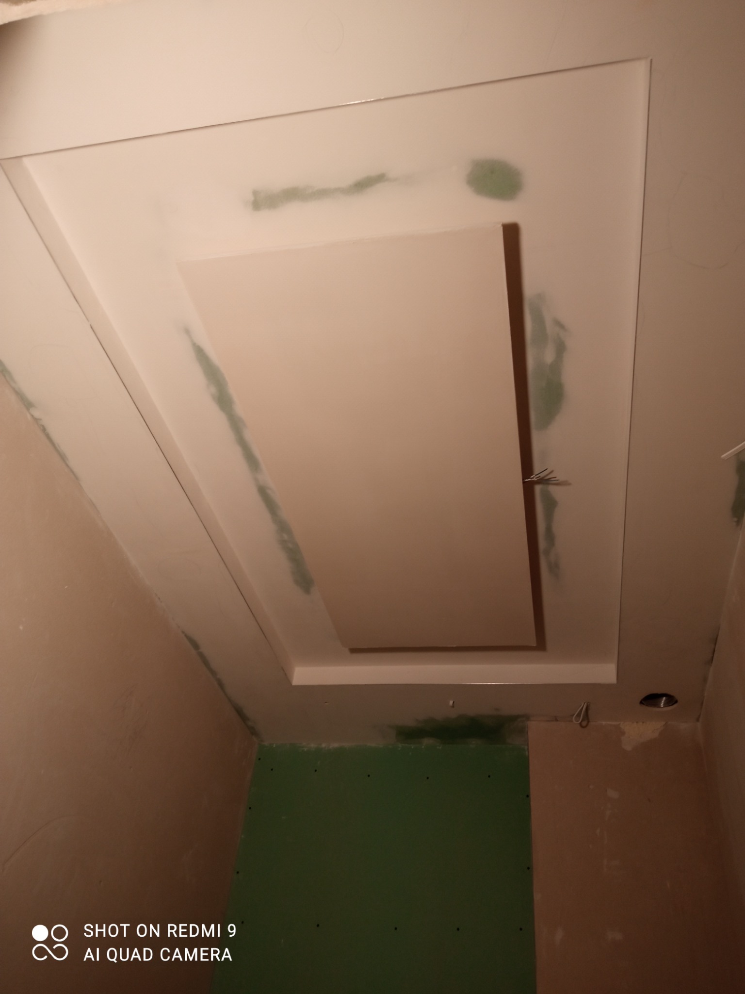 Sufit podwieszany z płyty kartonowo-gipsowej w trakcie montażu, widoczne ślady szpachlowania i łączenia płyt, częściowo zielona płyta na ścianie, surowe ściany w odcieniach beżu, kabel wystający...