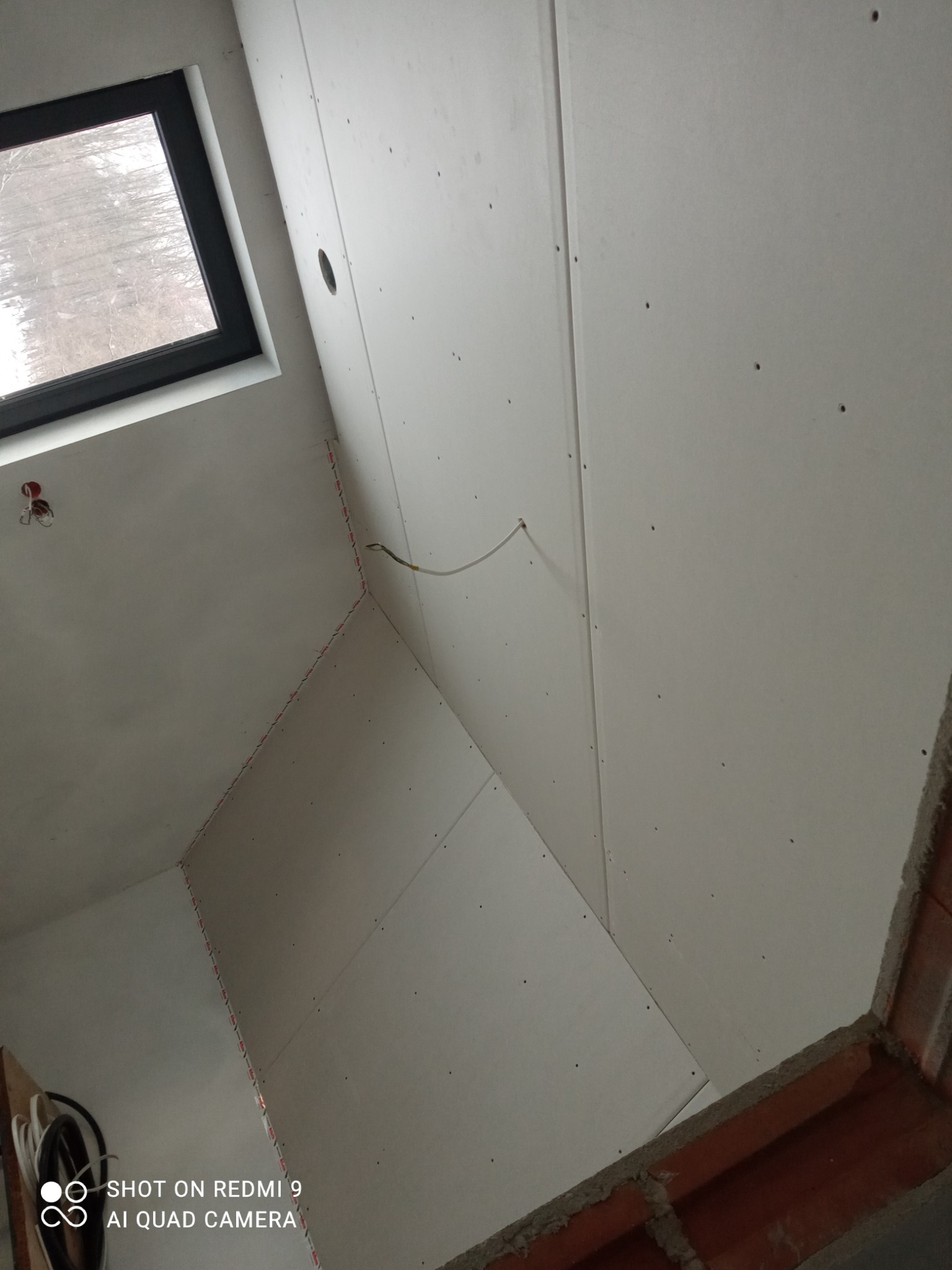 Skośny sufit poddasza z zamontowanymi białymi płytami, widoczne łączenia płyt i otwór na oświetlenie, okno dachowe w tle.