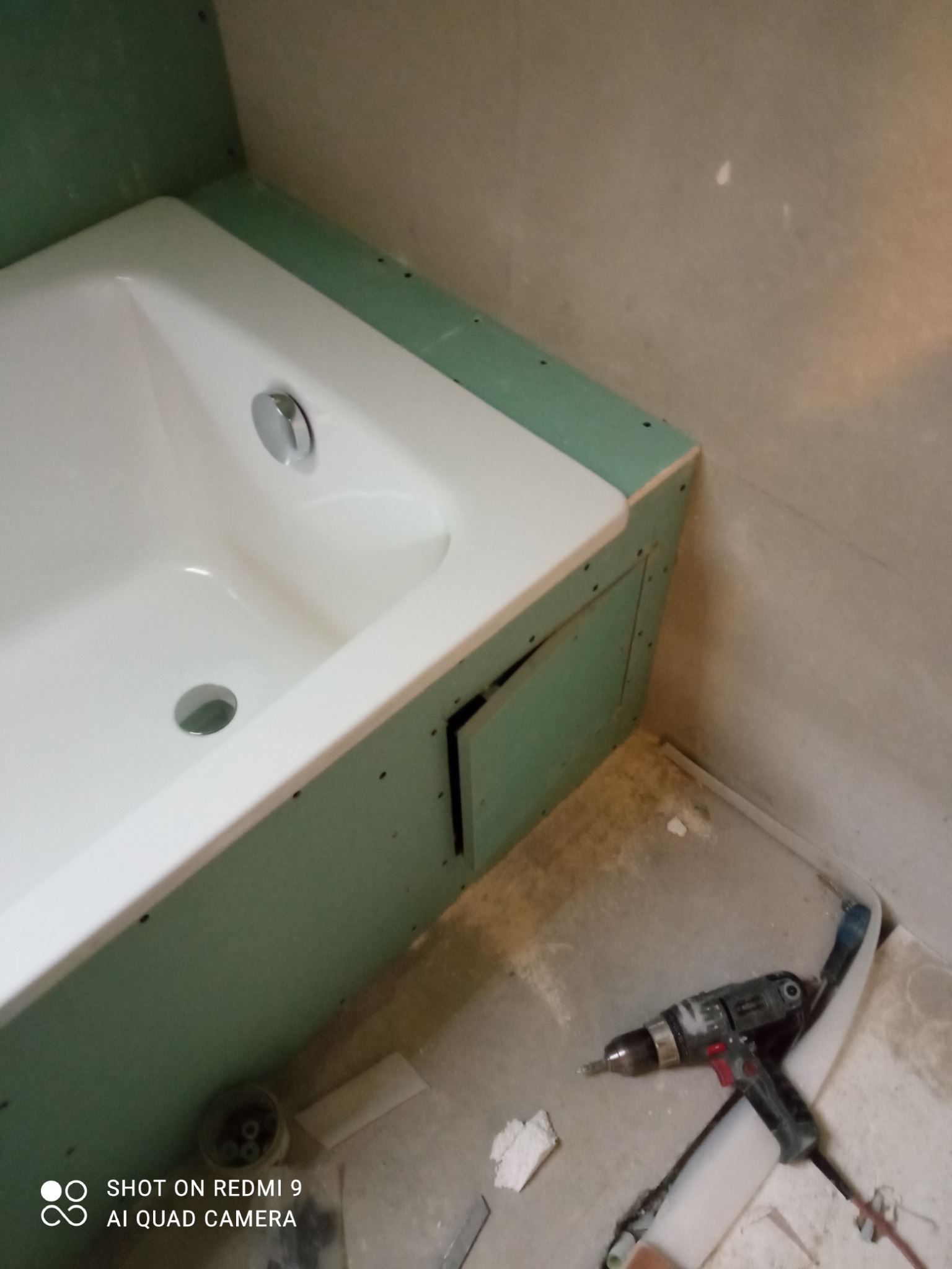 Biała wanna obudowana zielonymi płytami gipsowo-kartonowymi, widoczny otwór rewizyjny i wkrętarka leżąca na podłodze.