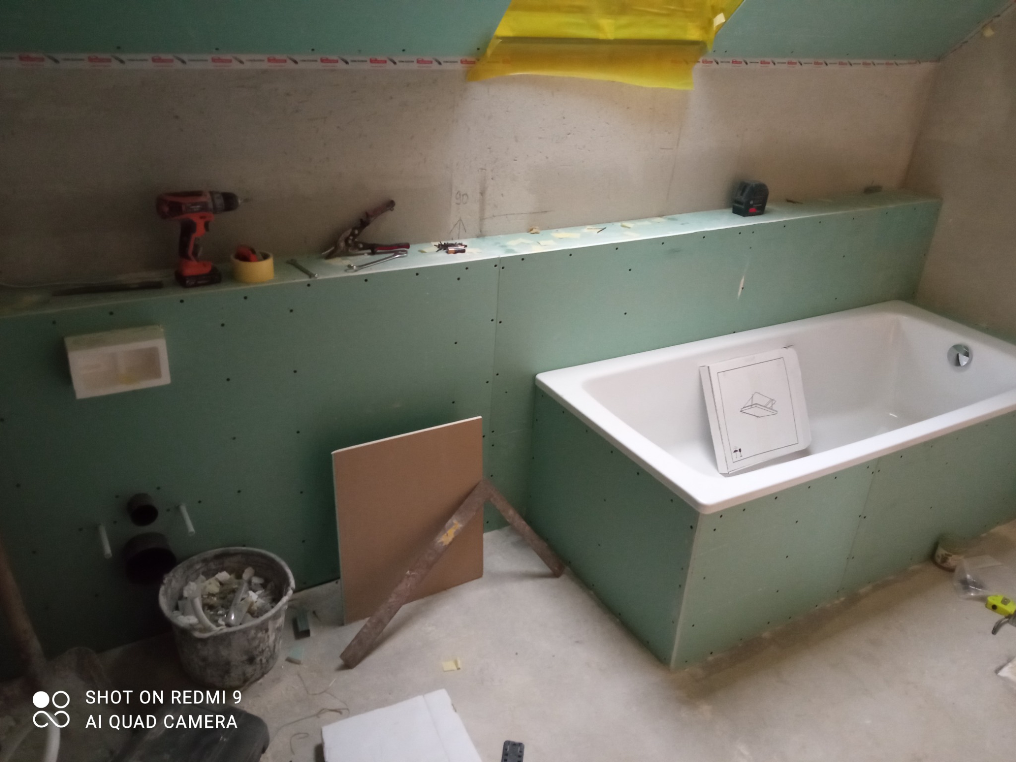 Łazienka w trakcie remontu: wanna obudowana zielonymi płytami, widoczne narzędzia, poziomica laserowa, wiadro z odpadami i karton z instrukcją montażu w wannie.