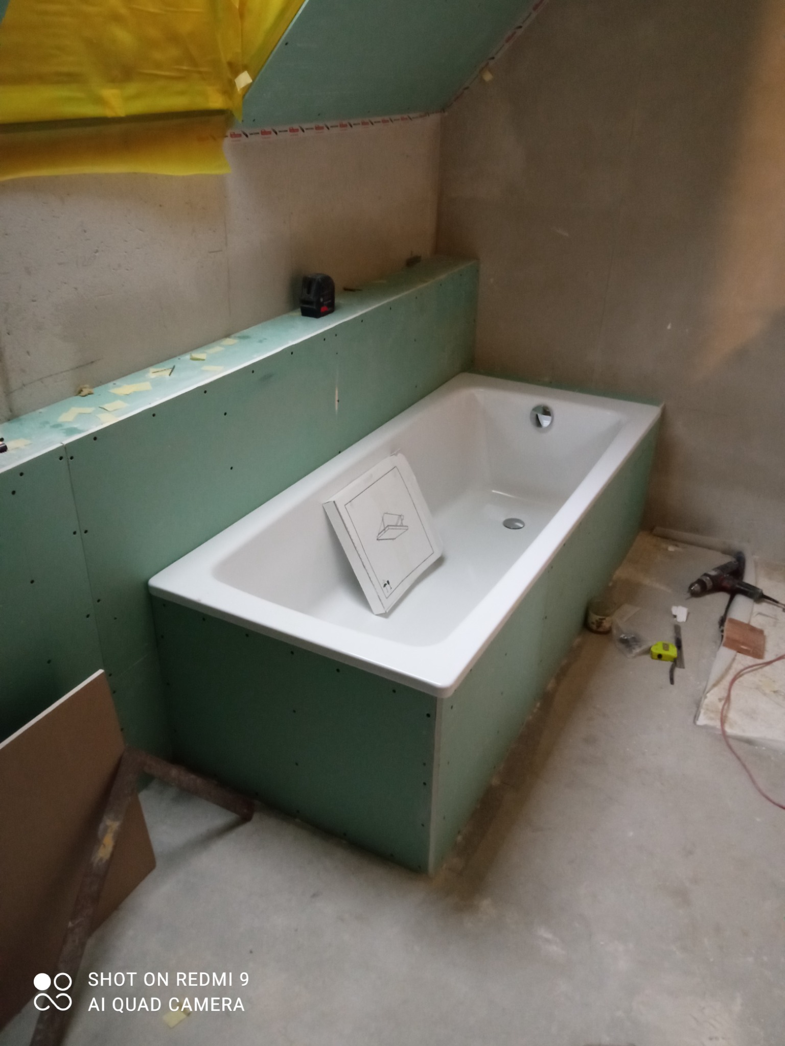 Biała wanna obudowana zieloną płytą, częściowo zamontowana w łazience na poddaszu. Na wannie leży instrukcja montażu. W tle widoczny poziomica laserowa i żółta folia zabezpieczająca.