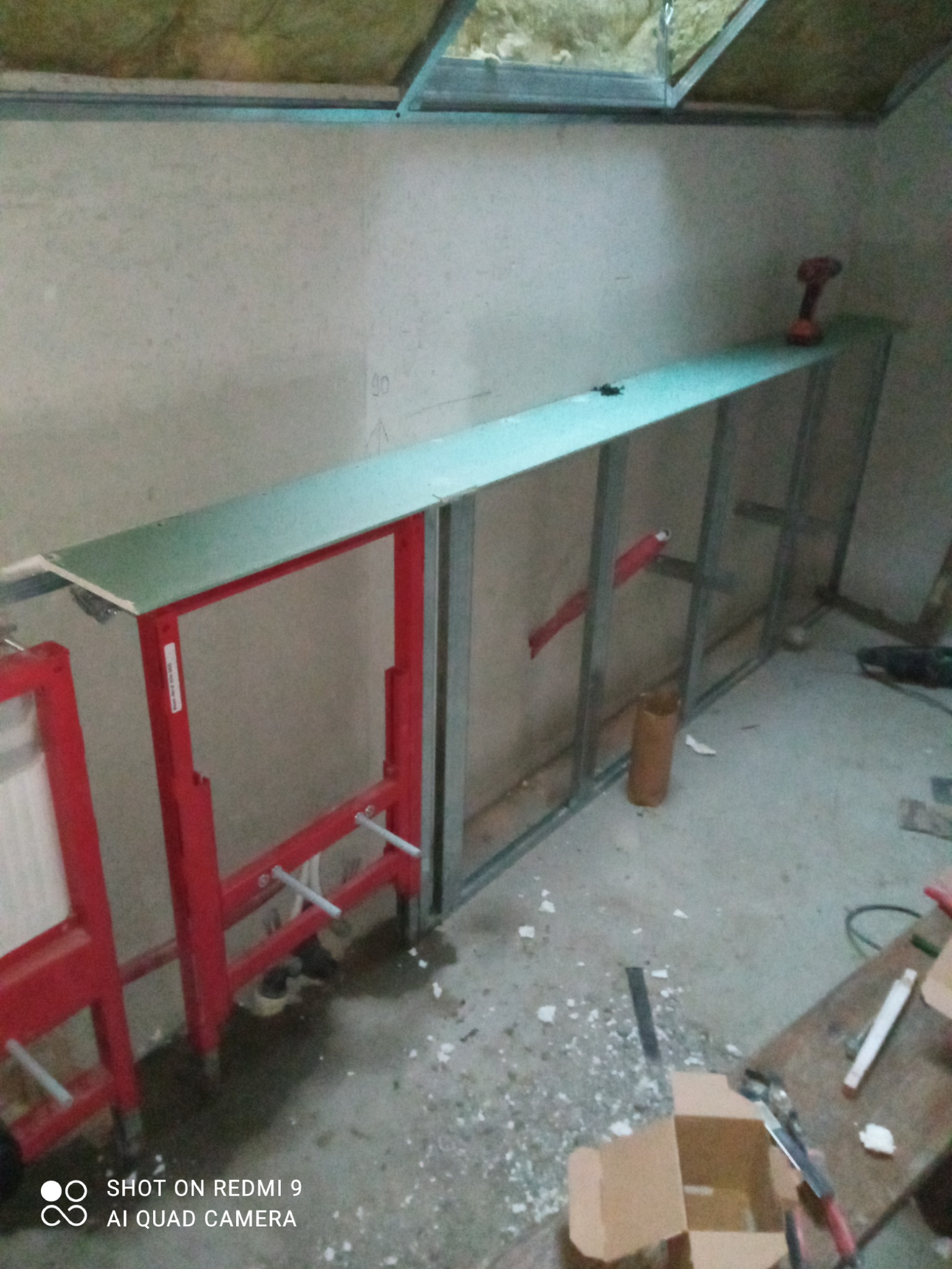 Wnętrze łazienki w trakcie remontu: widoczne metalowe profile konstrukcyjne, czerwona rama pod zabudowę WC, częściowo zamontowana zielona płyta na stelażu, w tle narzędzia i wkrętarka na płycie.