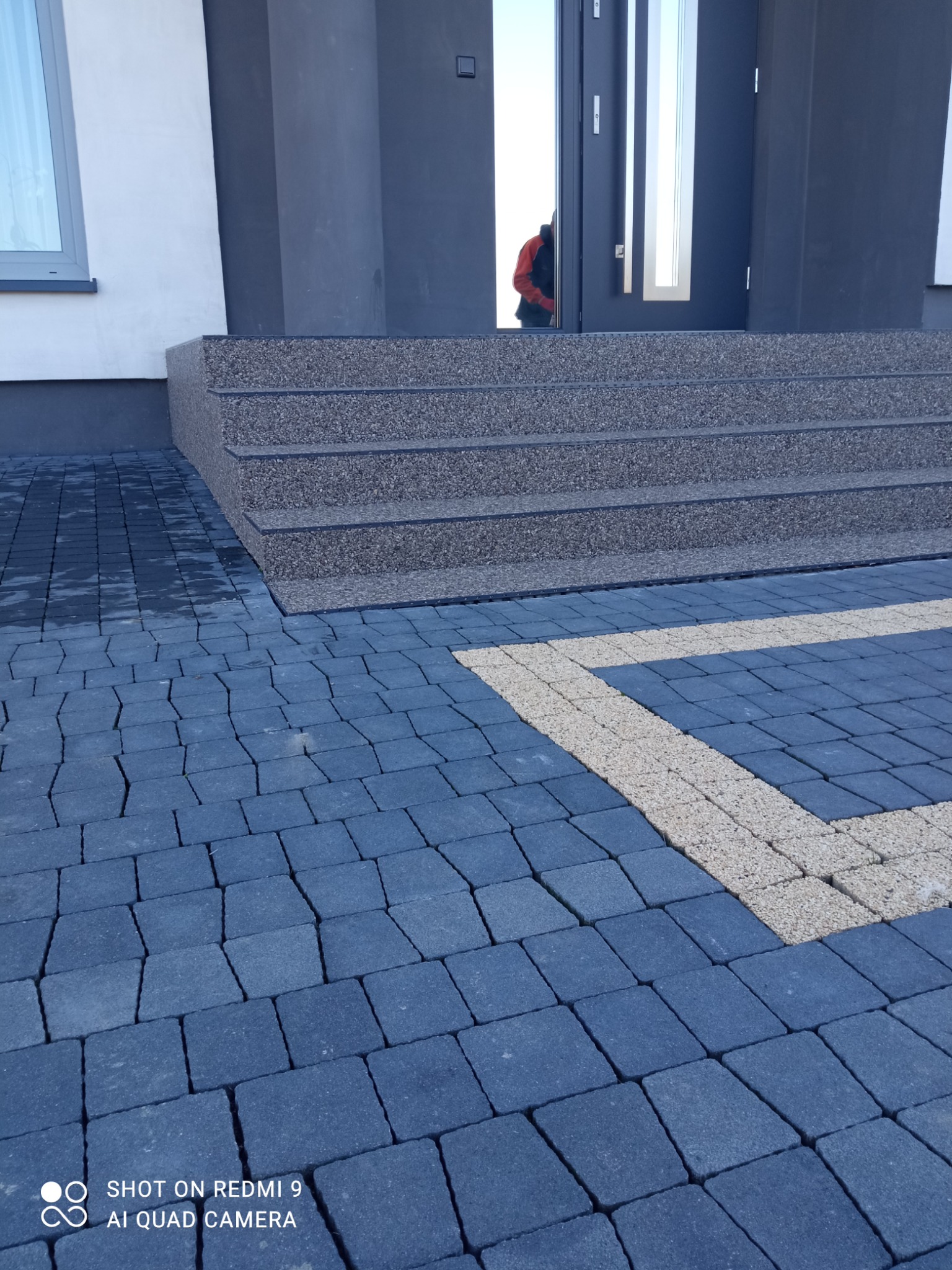 Ułożona kostka brukowa w odcieniach szarości i beżu przed wejściem do budynku z trzema schodami pokrytymi kruszywem.