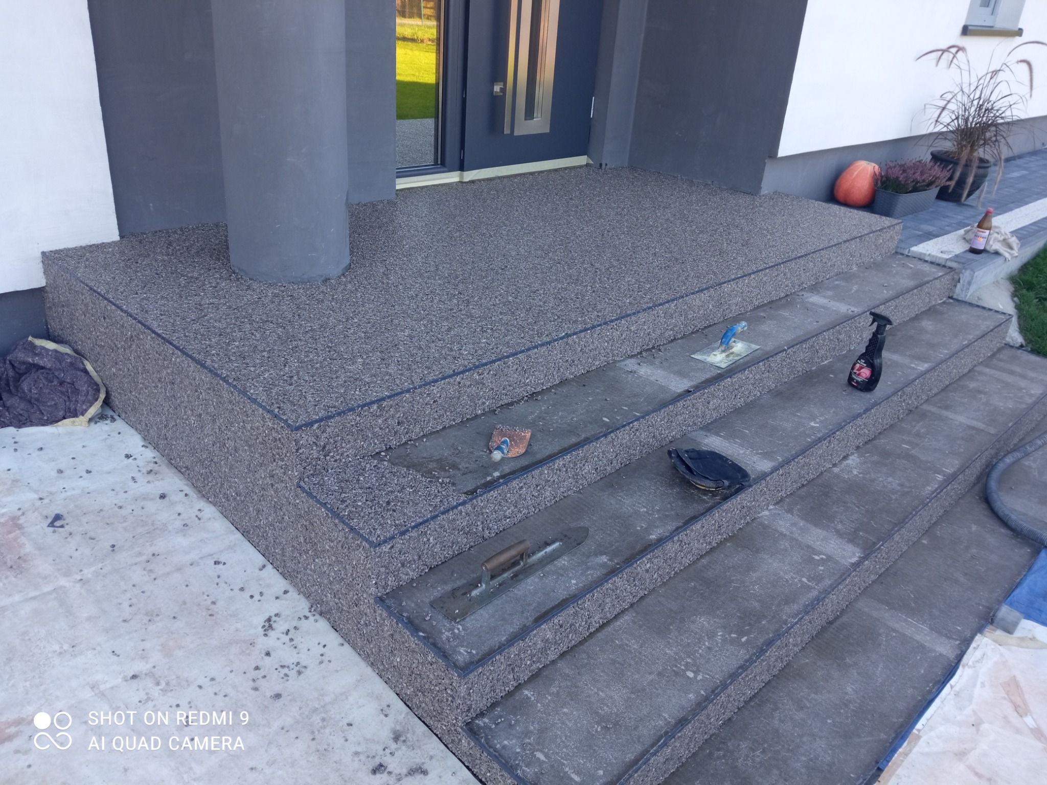 Schody wejściowe do domu wykończone szarą, kamyczkową posadzką żywiczną, widoczne narzędzia pracy i butelka z płynem czyszczącym.
