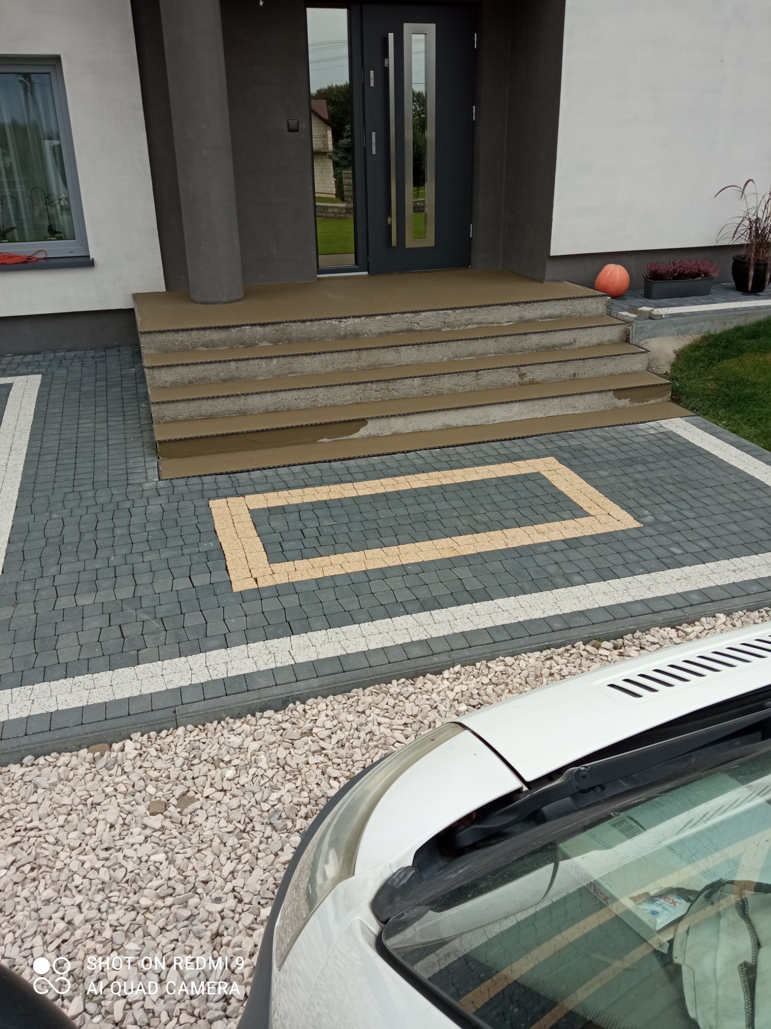 Ułożona kostka brukowa w szaro-biało-żółte wzory przed wejściem do domu z trzema schodami, widoczny fragment białego samochodu.