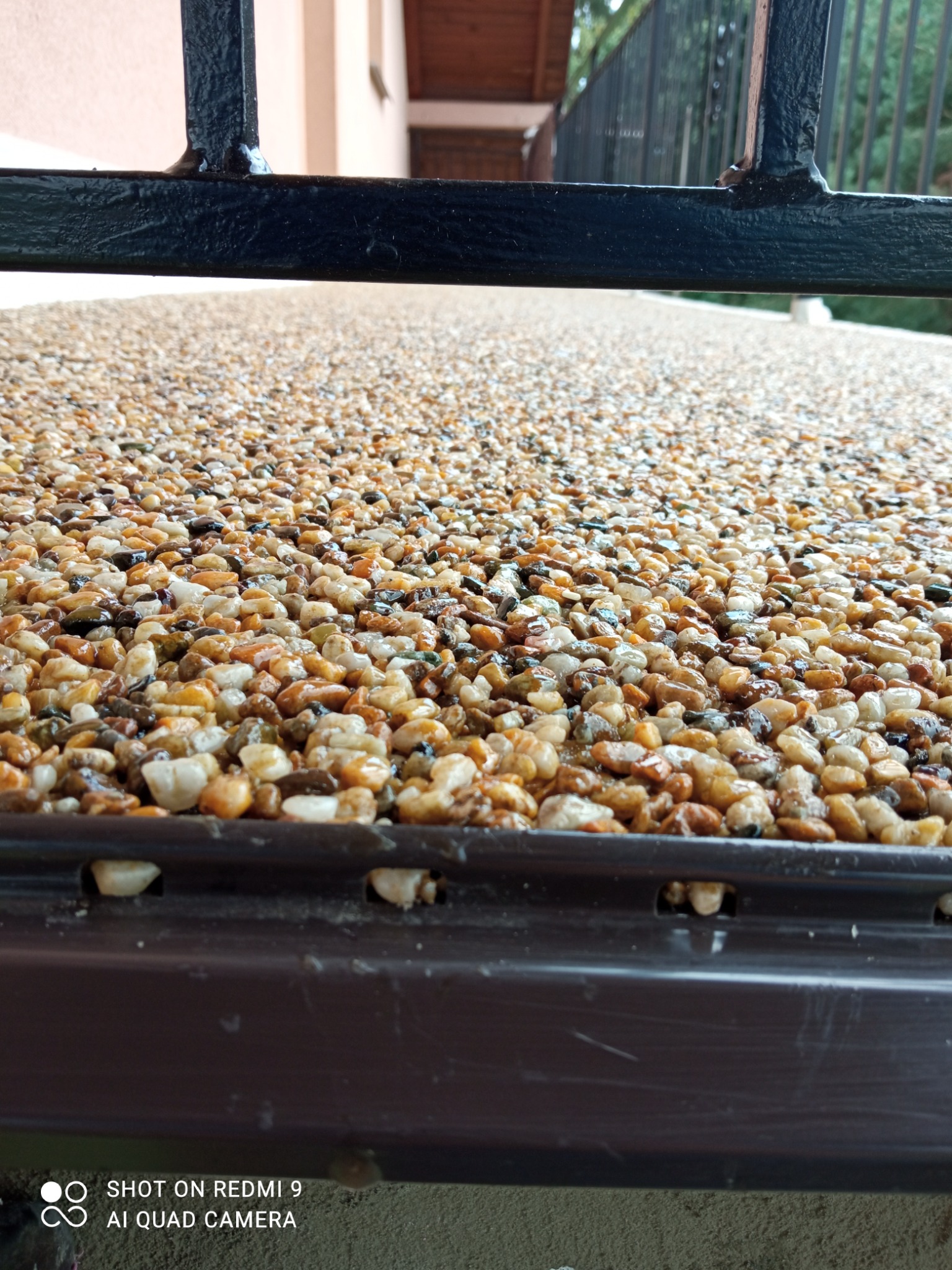 Bursztynowa posadzka z żywicy epoksydowej, widok z poziomu gruntu przez ozdobne metalowe ogrodzenie, ukazujący drobne kamyczki zatopione w błyszczącej powierzchni.