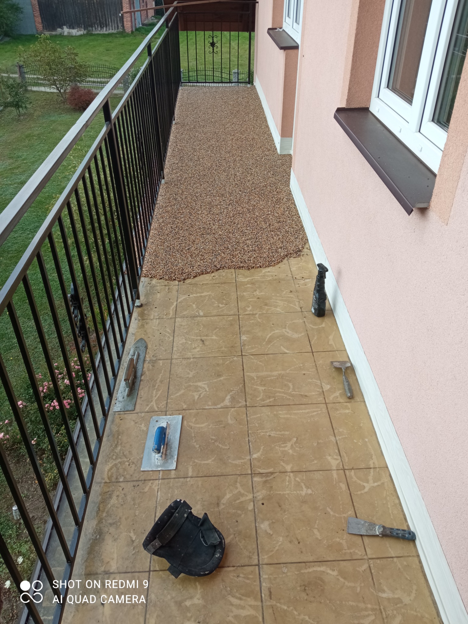 Balkon w trakcie wykańczania z częściowo nałożoną posadzką żywiczną, widoczne narzędzia: szpachelki, kielnie i spryskiwacz, brązowe płytki pod spodem.