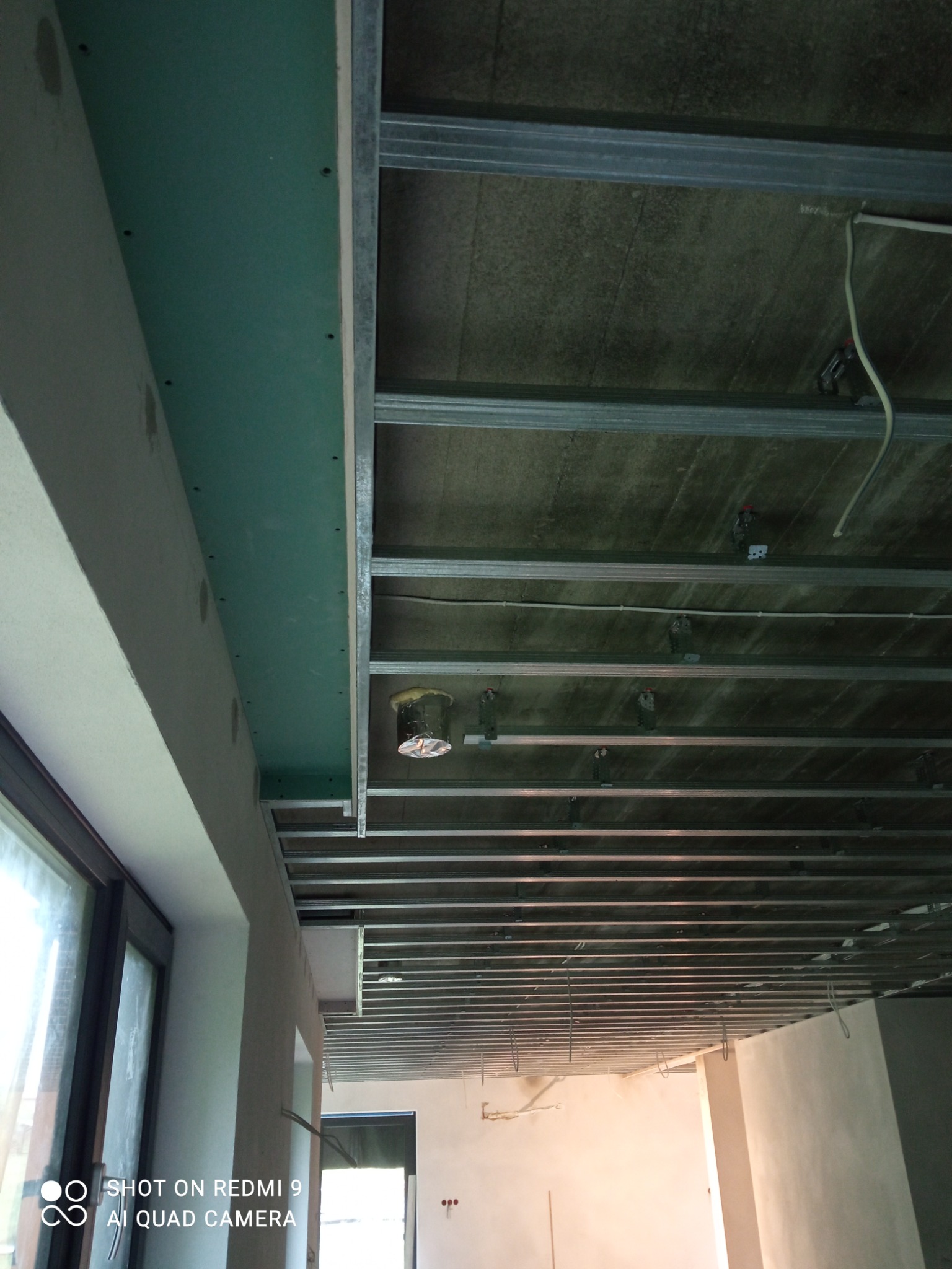 Montaż sufitu podwieszanego z płyt karton gips, widoczne profile metalowe i częściowo zamontowana zielona płyta GK przy oknie.