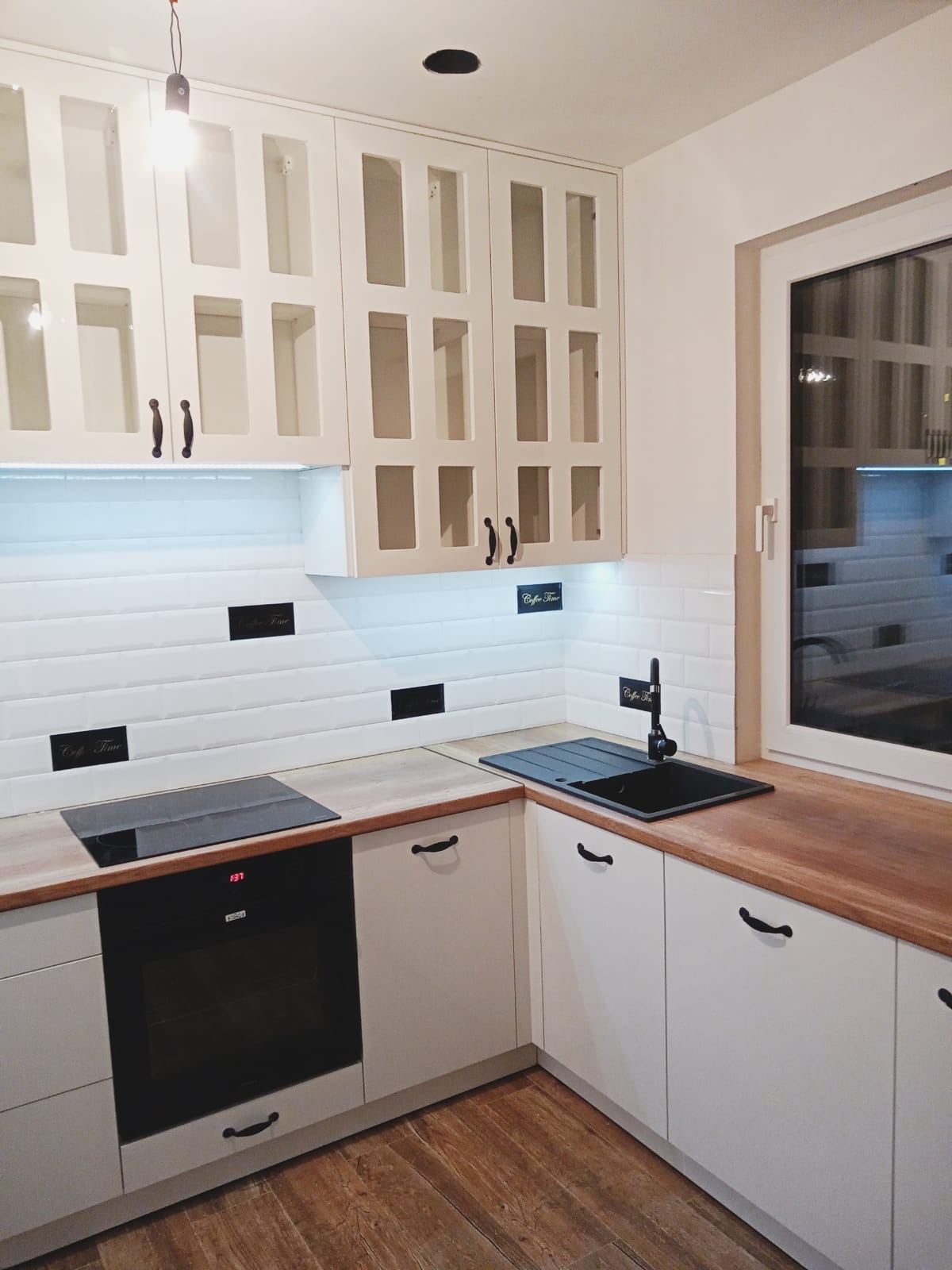 Jasna kuchnia z białymi szafkami z przeszklonymi frontami, drewnianym blatem i czarnymi akcentami, takimi jak bateria i płyta grzewcza.