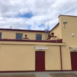 Żółty budynek z czerwonymi akcentami, widoczne okna z roletami, drzwi wejściowe z daszkami, drabina na dachu i elewacji, oraz lampa zewnętrzna.