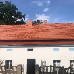 Dwupiętrowy dom z nowym, czerwonym dachem z dachówki ceramicznej, dwoma kominami z cegły i białymi ścianami z niebieskimi akcentami, widoczna drabina przy wejściu, ogrodzenie z betonowymi słupkami,...