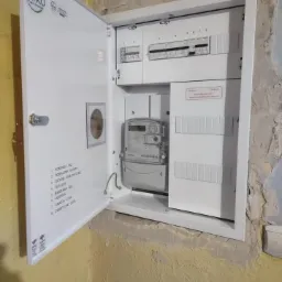 Otwarta, biała skrzynka elektryczna z licznikiem i bezpiecznikami, zamontowana na żółtej, nierównej ścianie. Na drzwiach skrzynki widoczna naklejka z opisem elementów i logo firmy FRIKO.