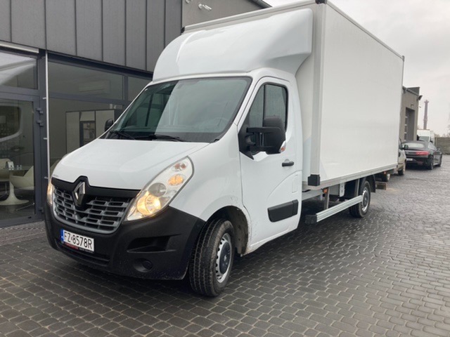 Biały Renault Master z zabudową kontenerową, zaparkowany na kostce brukowej przed budynkiem z przeszklonym wejściem, widoczne tablice rejestracyjne.