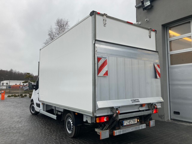 Tył białego samochodu dostawczego z zabudową kontenerową, widoczne światła, tablica rejestracyjna i oznaczenia ostrzegawcze, na tle szarego budynku z bramą garażową.