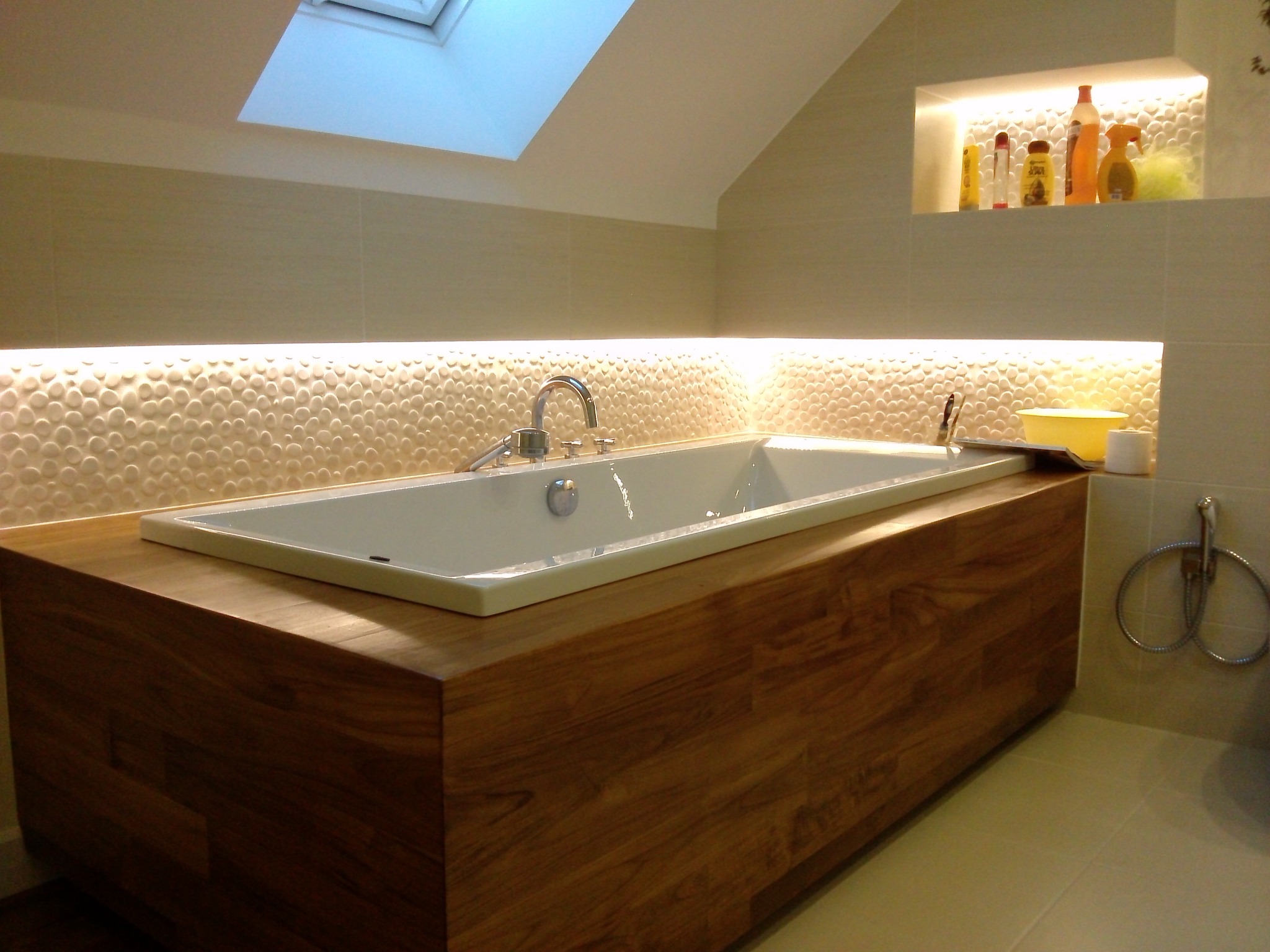 Nowoczesna łazienka na poddaszu z wanną obudowaną drewnem, oświetleniem LED i dekoracyjną ścianą z wypukłymi okrągłymi płytkami, widok na okno dachowe.