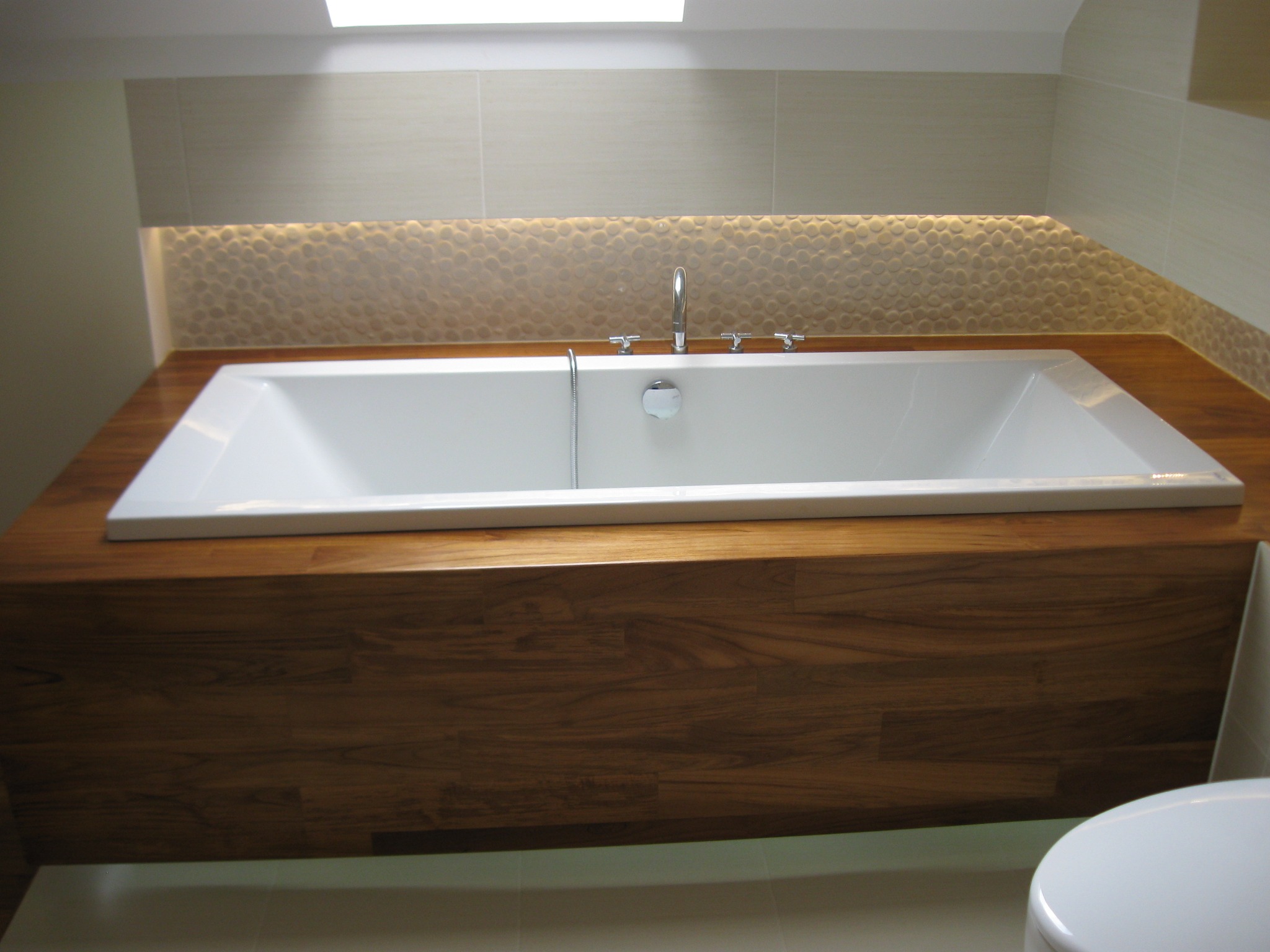 Nowoczesna, podświetlona wanna w zabudowie z drewna egzotycznego, z baterią czterootworową i mozaiką na ścianie w tle.