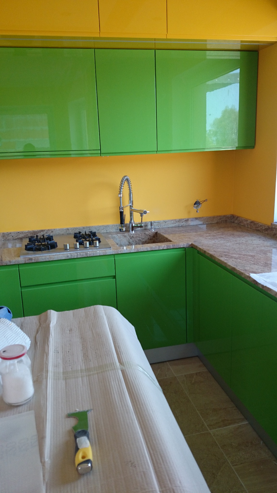 Montaż zielonych szafek kuchennych z żółtymi akcentami w Warszawie, widoczna płyta grzewcza i zlew, blat z kamienia oraz narzędzie do wygładzania na zabezpieczonej powierzchni.