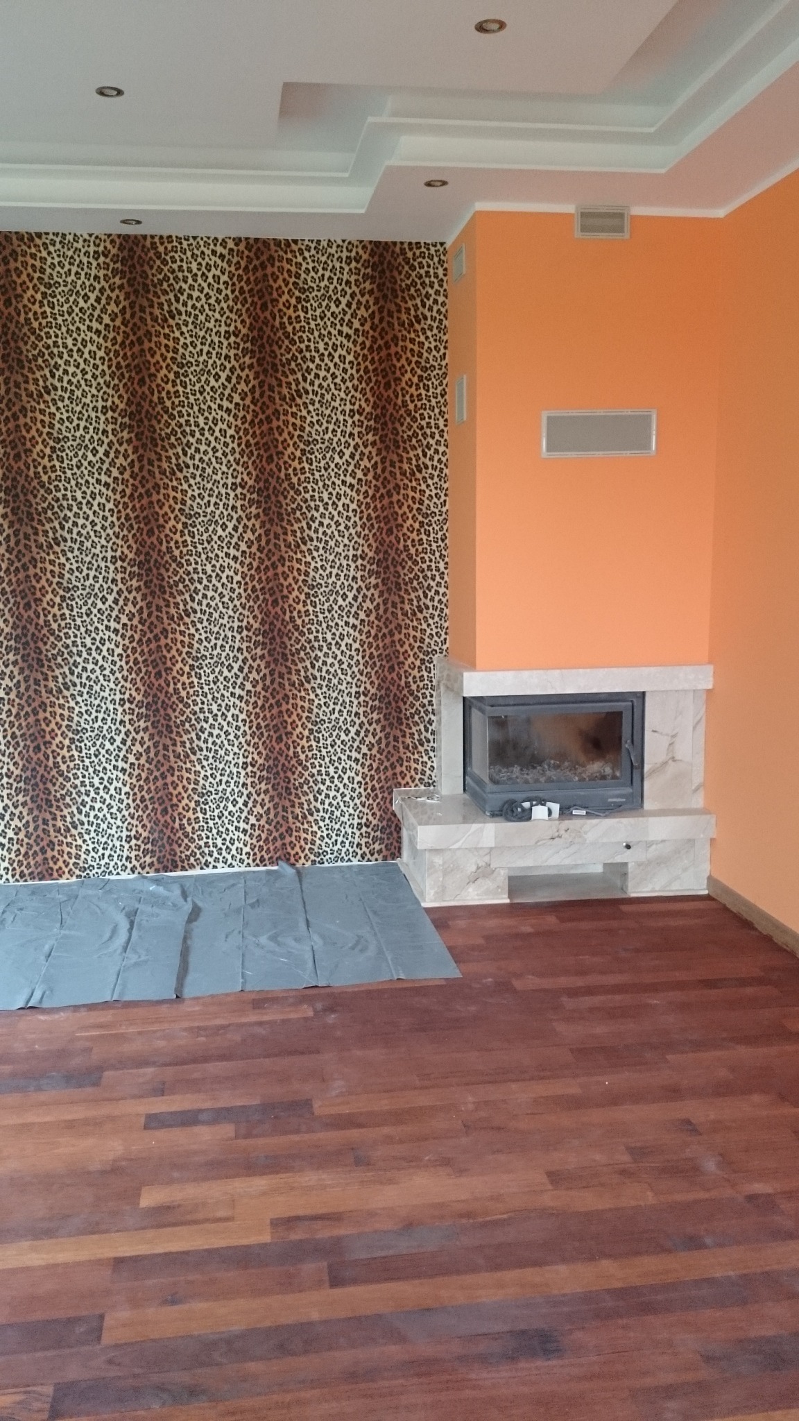 Pokój po remoncie z drewnianą podłogą, ścianą w panterkę i marmurowym kominkiem, zabezpieczony folią malarską.
