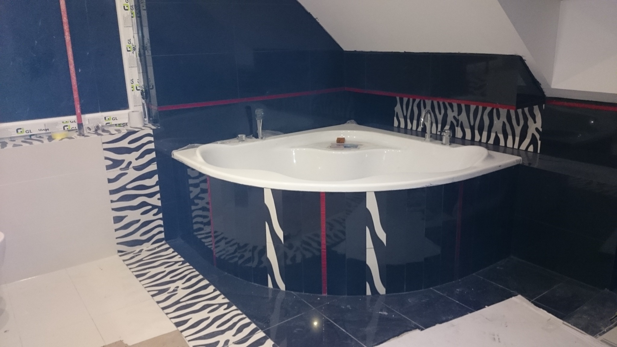 Biała, narożna wanna w łazience z czarnymi płytkami, akcentami w kolorze czerwonym i wzorem zebry na ścianach i podłodze.