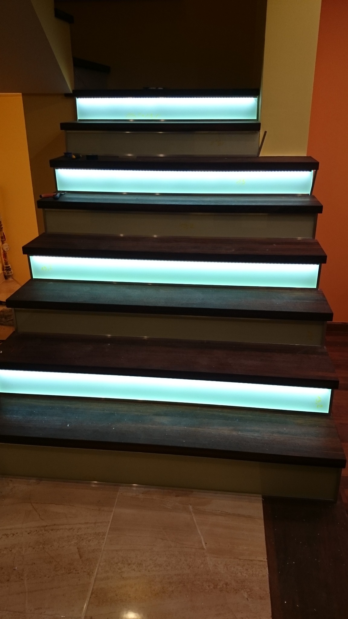 Schody z drewnianymi stopniami podświetlonymi od spodu turkusowymi ledami, widok z dołu na kilka stopni, ściany w odcieniach żółci i pomarańczy, podłoga wyłożona płytkami imitującymi marmur.