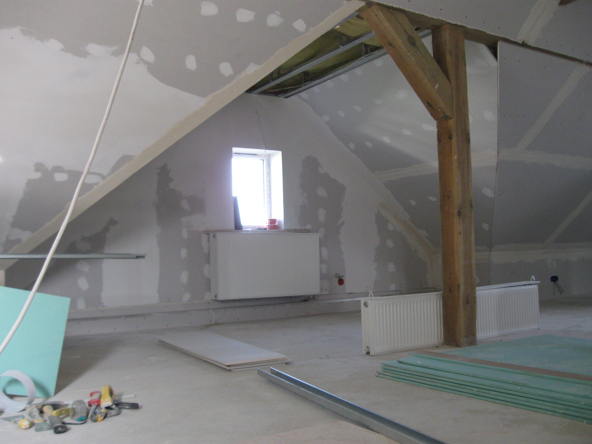 Adaptacja poddasza: surowe ściany z płyt gipsowo-kartonowych, drewniana belka konstrukcyjna, grzejniki i narzędzia na podłodze, widoczne ślady szpachlowania.