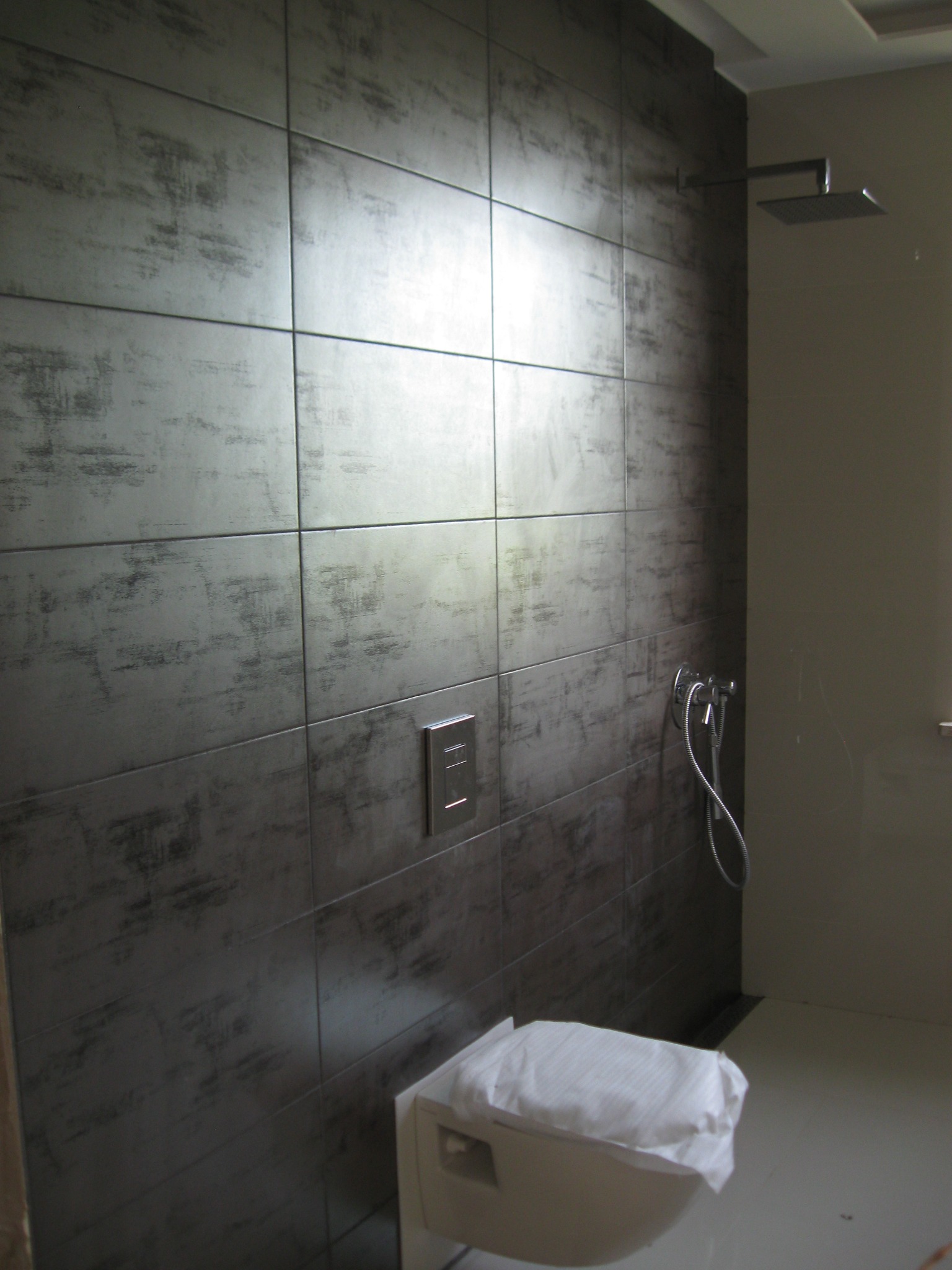 Nowoczesna łazienka z ciemnoszarą, dużą płytką imitującą beton na ścianach, chromowaną armaturą prysznicową i podtynkową spłuczką WC z białą deską zabezpieczoną folią.