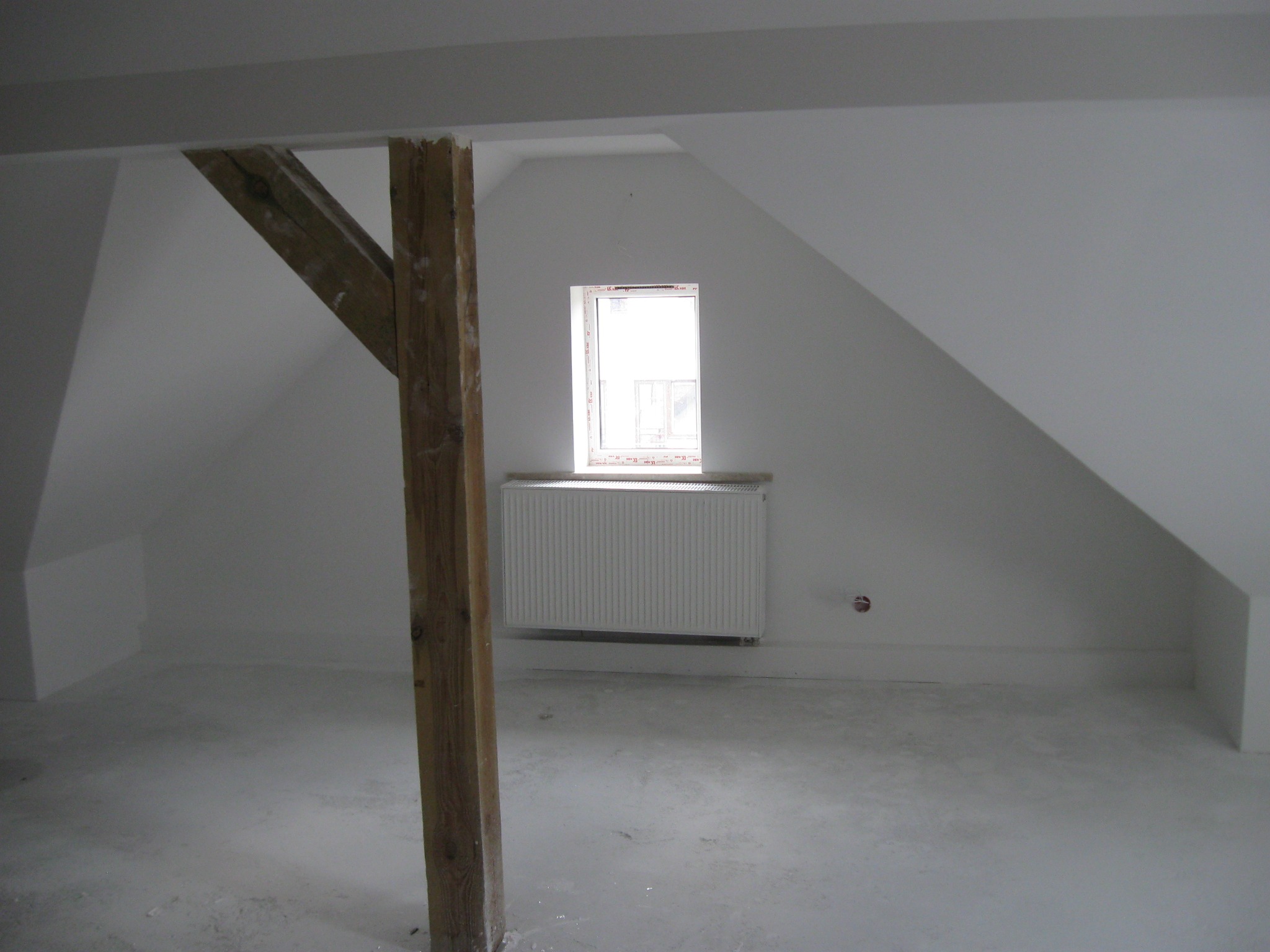Poddasze po wylewce, widoczny drewniany słup konstrukcyjny, okno i grzejnik na tle białych ścian ze skosami.