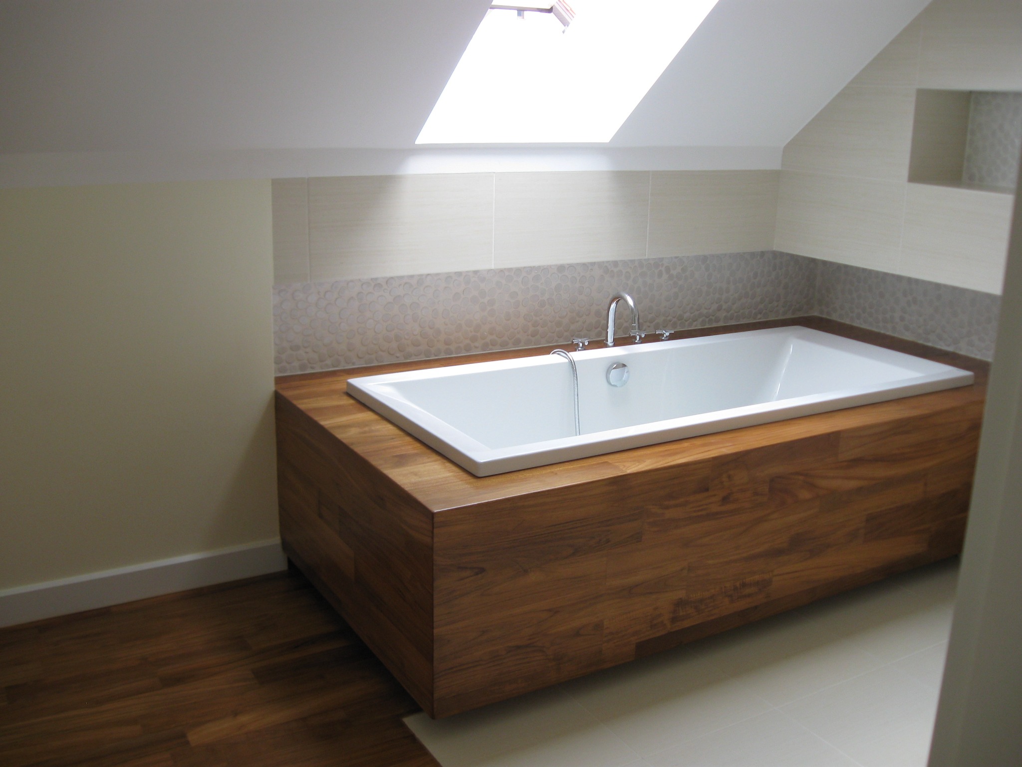 Biała wanna w drewnianej obudowie w łazience na poddaszu z oknem dachowym, jasnymi płytkami i drewnianą podłogą.