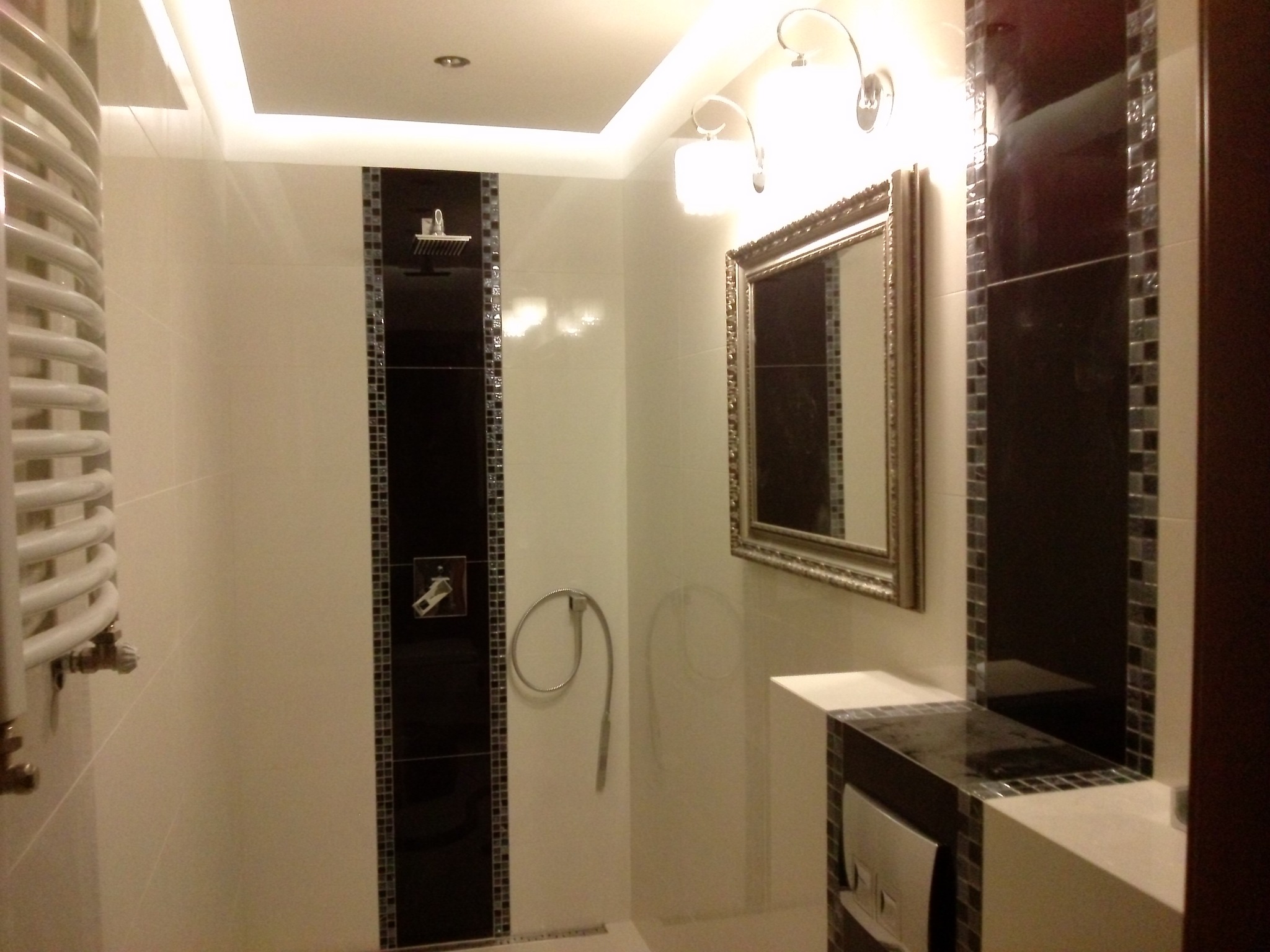 Nowoczesna łazienka z czarno-białymi płytkami mozaikowymi, grzejnikiem drabinkowym, lustrem w ozdobnej ramie i oświetleniem punktowym.