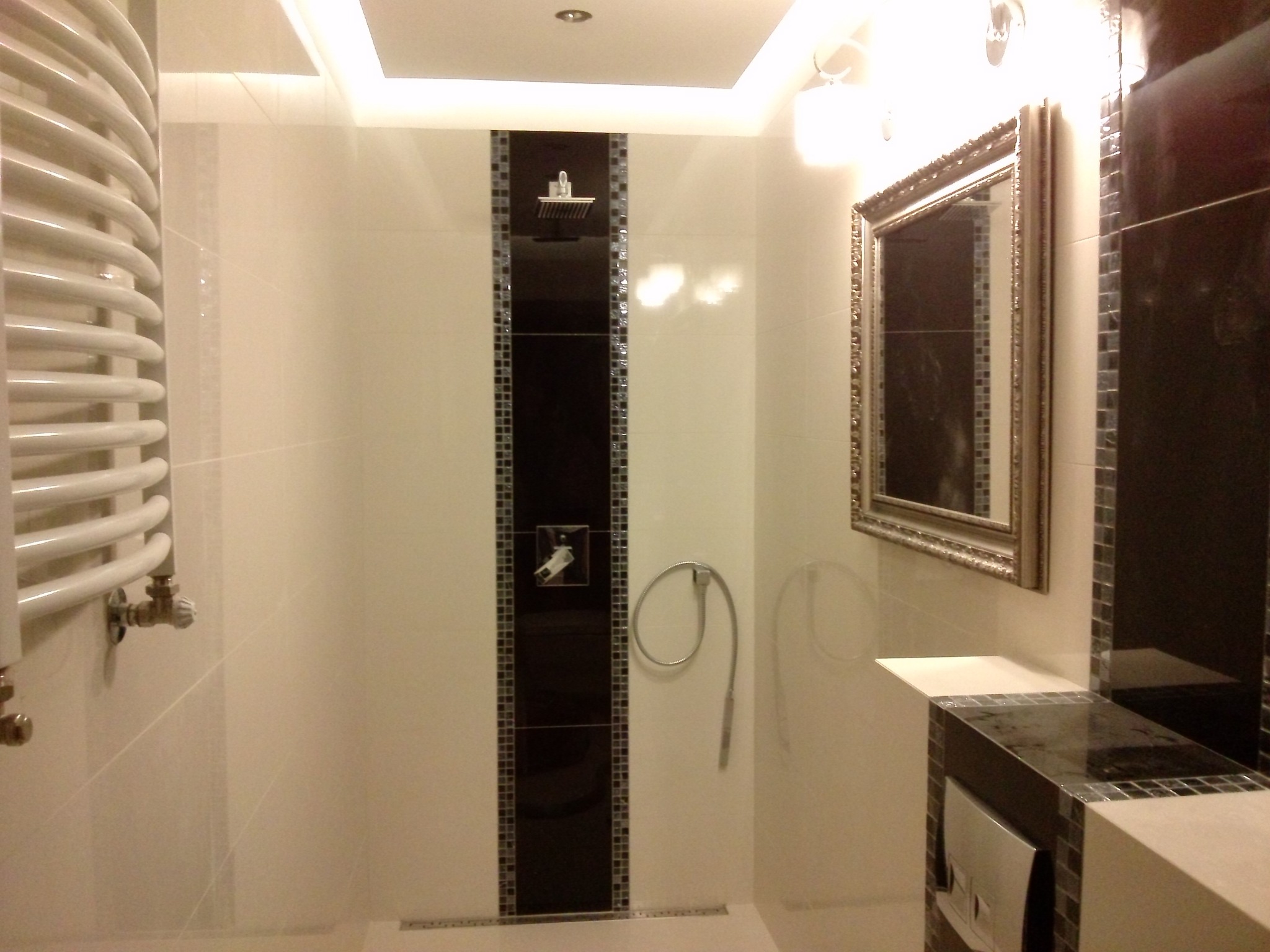 Nowoczesna łazienka z białymi i czarnymi płytkami, grzejnikiem drabinkowym, lustrem w ozdobnej ramie i deszczownicą wkomponowaną w czarny pas płytek.