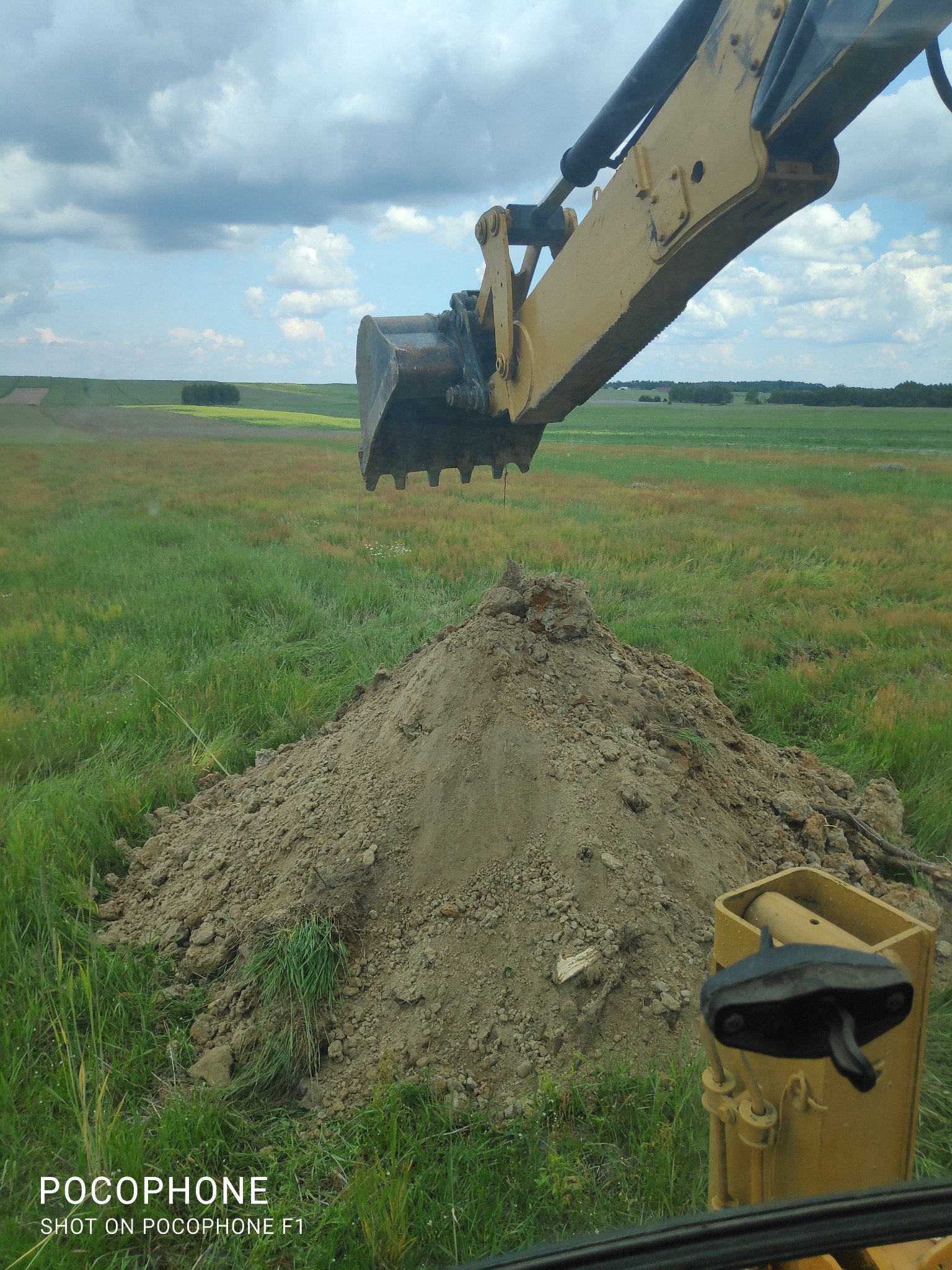 Żółta koparka z łyżką nad stertą ziemi na zarośniętym polu, widok z kabiny operatora, w tle zielone pola i pochmurne niebo.
