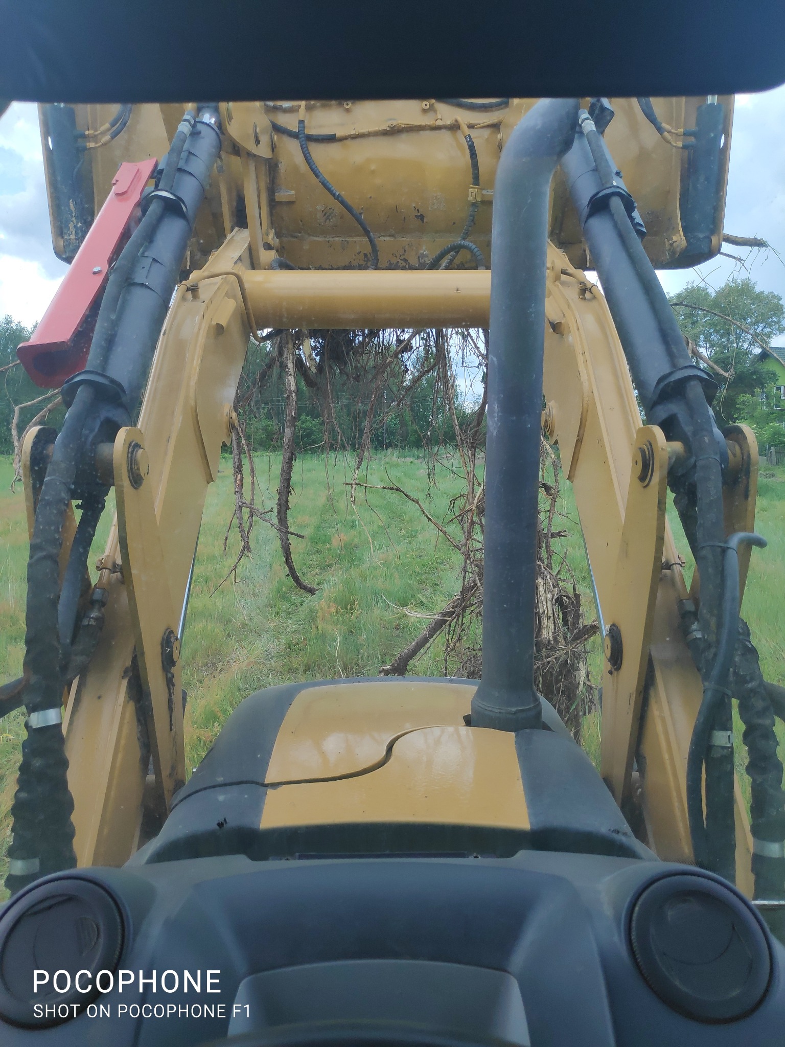 Widok z kabiny koparko-ładowarki na uniesione ramię z korzeniami i ziemią, w tle zielona łąka i zabudowania.
