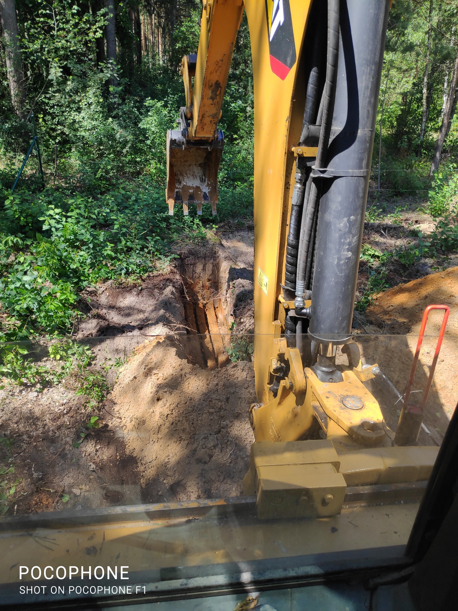Żółta koparka z łyżką w wykopie, widok z kabiny operatora, las w tle.