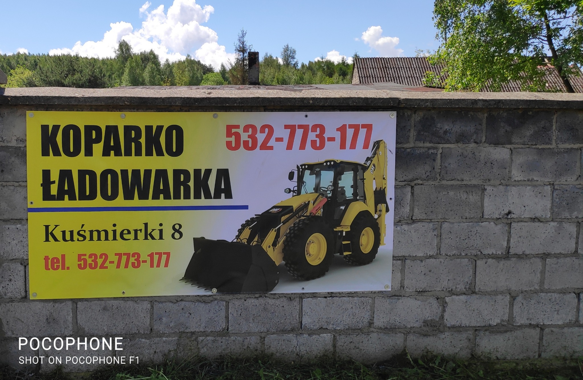Ogłoszenie na betonowym murze reklamujące usługi koparko-ładowarki z numerem telefonu i adresem, z ilustracją żółtej maszyny budowlanej CAT.