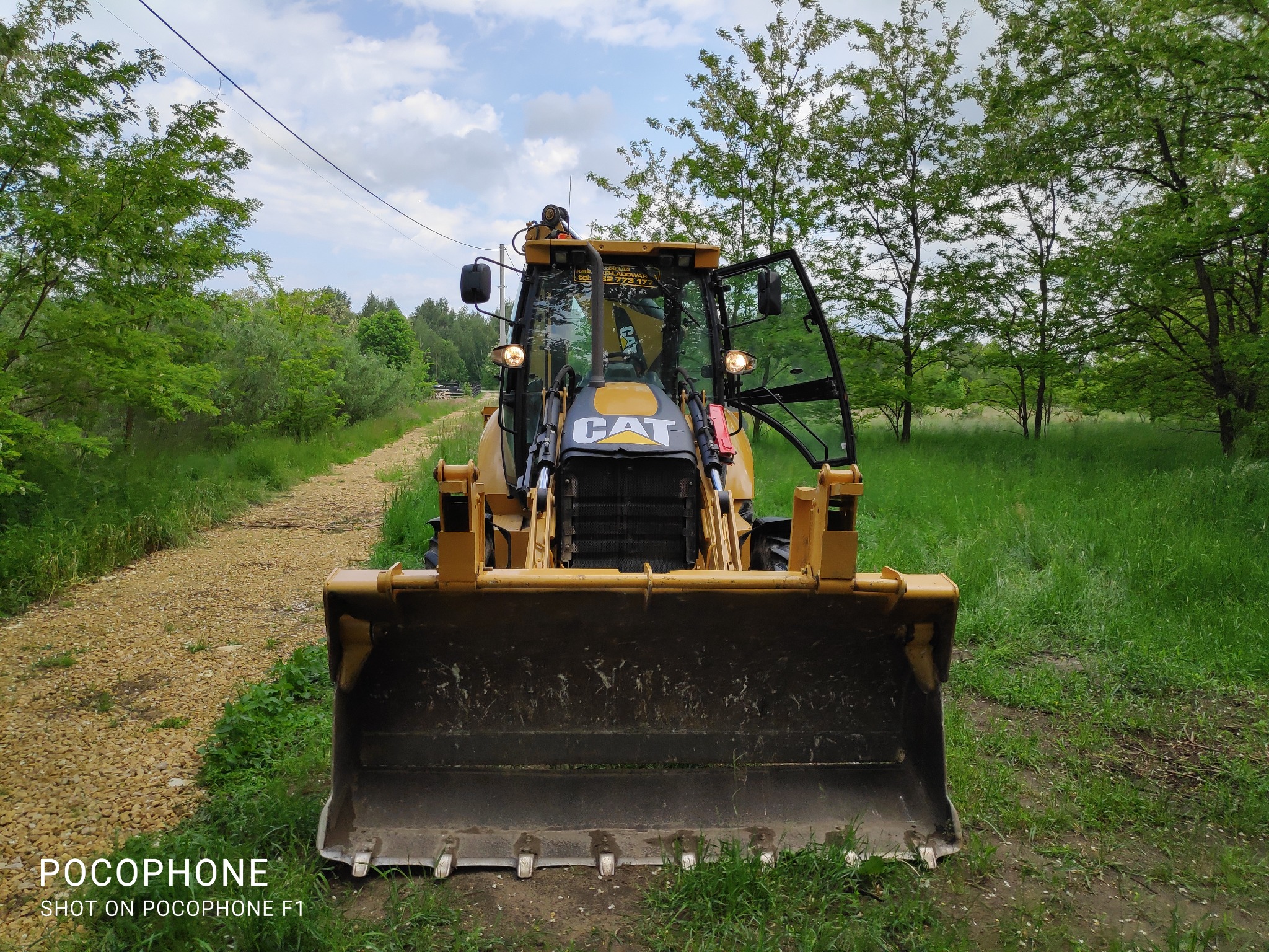 Żółta koparko-ładowarka CAT stojąca na polnej drodze, z otwartymi drzwiami kabiny, widok z przodu na łyżkę, w tle zielona roślinność i częściowo zachmurzone niebo.