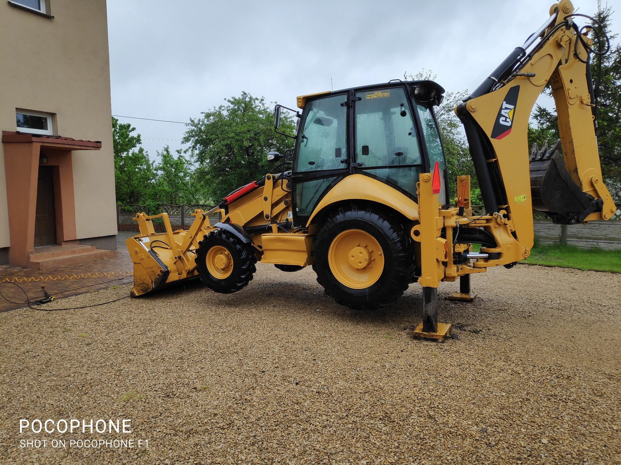 Żółta koparko-ładowarka Cat 432E zaparkowana na żwirowym podłożu przed budynkiem i zielenią, widoczna z boku, z podpórkami opuszczonymi.