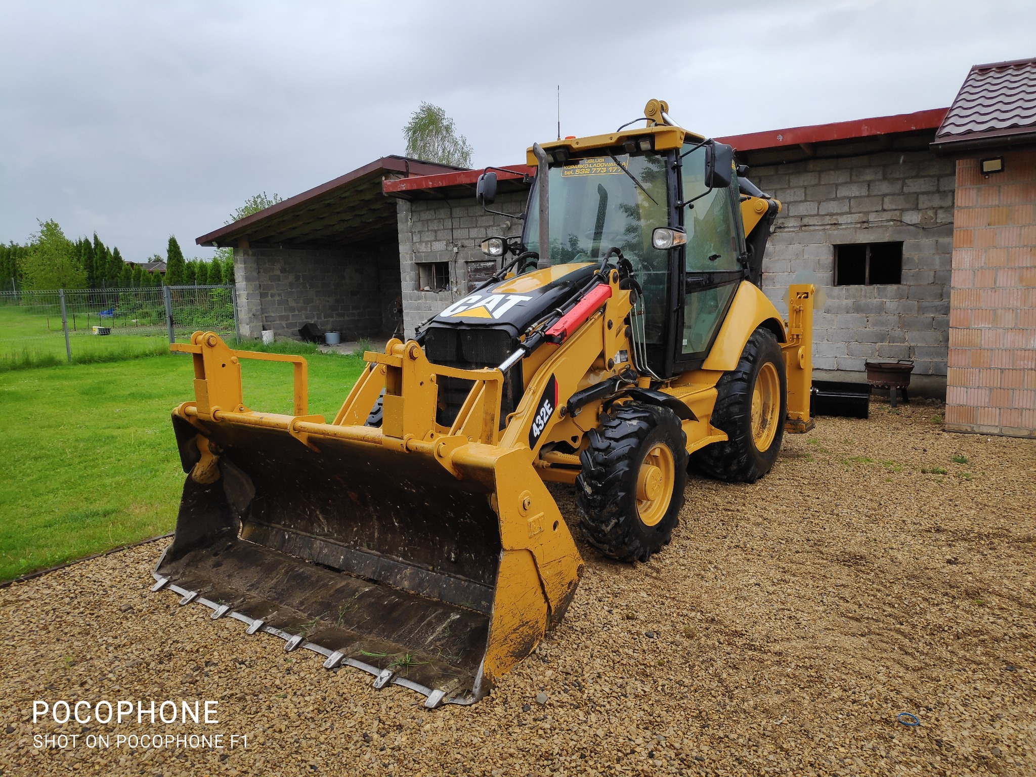 Żółta koparko-ładowarka CAT 432E zaparkowana na żwirowym podłożu przed budynkiem z szarego pustaka w pochmurny dzień.