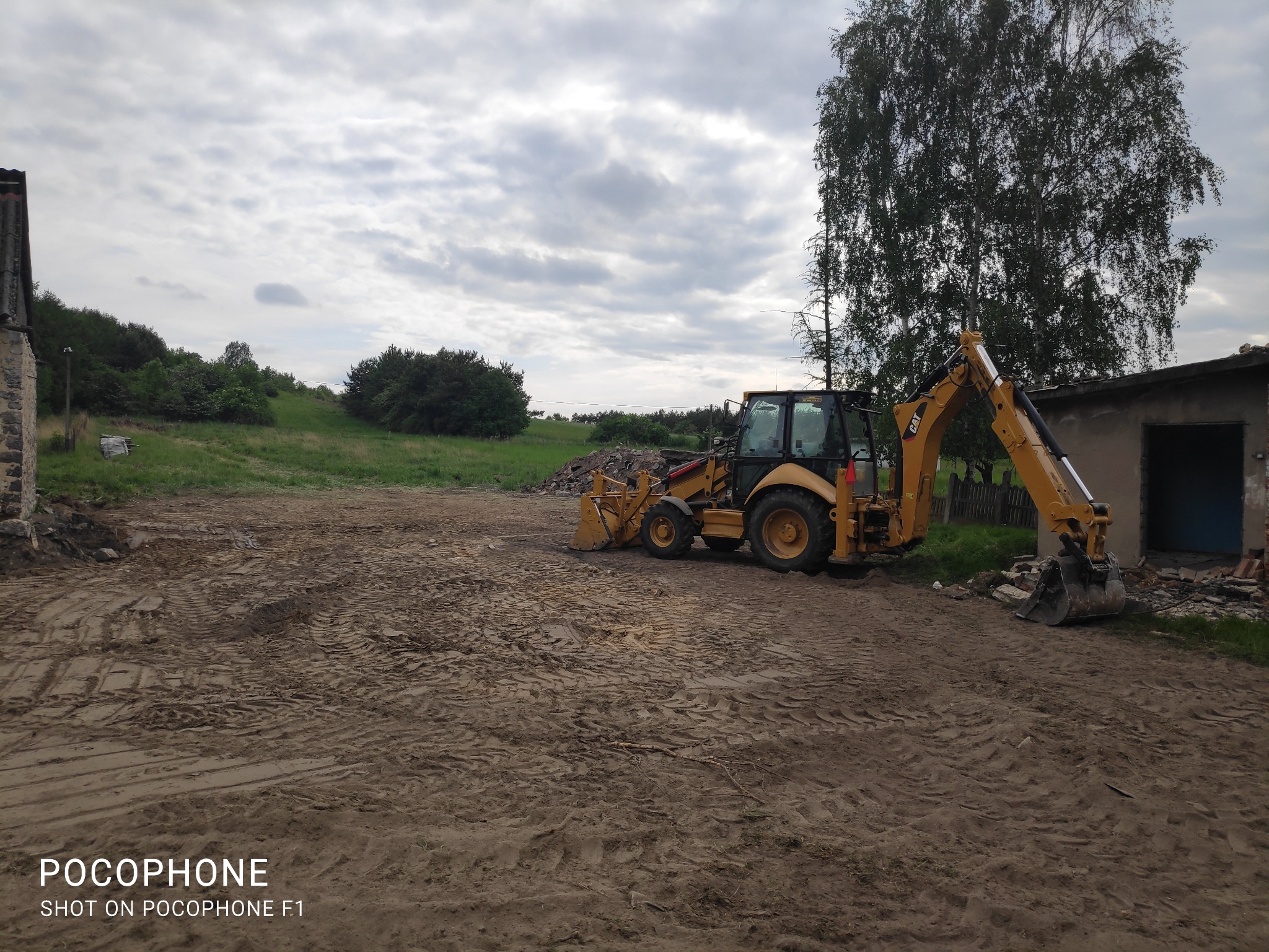 Żółta koparko-ładowarka CAT na placu budowy z gruzem, w tle zielone wzgórze pod pochmurnym niebem.