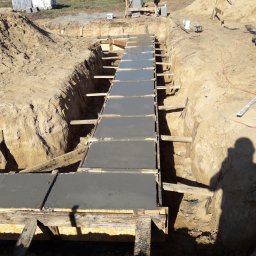 Usługi Remontowo-Budowlane - Świeżo wylany beton w szalunku fundamentów w wykopie, widoczne drewniane podpory i ziemia, na drugim planie budowa w toku i pracownik w ciemnej odzieży.