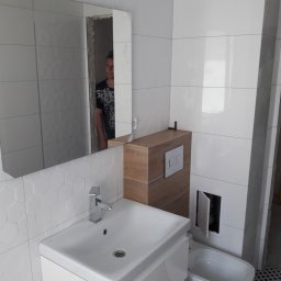 Usługi Remontowo-Budowlane - Nowoczesna łazienka w trakcie wykańczania: białe heksagonalne płytki ścienne, prostokątne białe płytki, umywalka z chromowaną baterią, zabudowa toalety z drewnopodobnym wykończeniem, odbicie osoby...