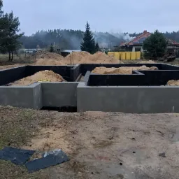 Wylane fundamenty domu jednorodzinnego, zabezpieczone czarną izolacją, z widocznymi prętami zbrojeniowymi i hałdami piasku, w tle dom z panelami słonecznymi i komin dymiący z sąsiedztwa lasu.