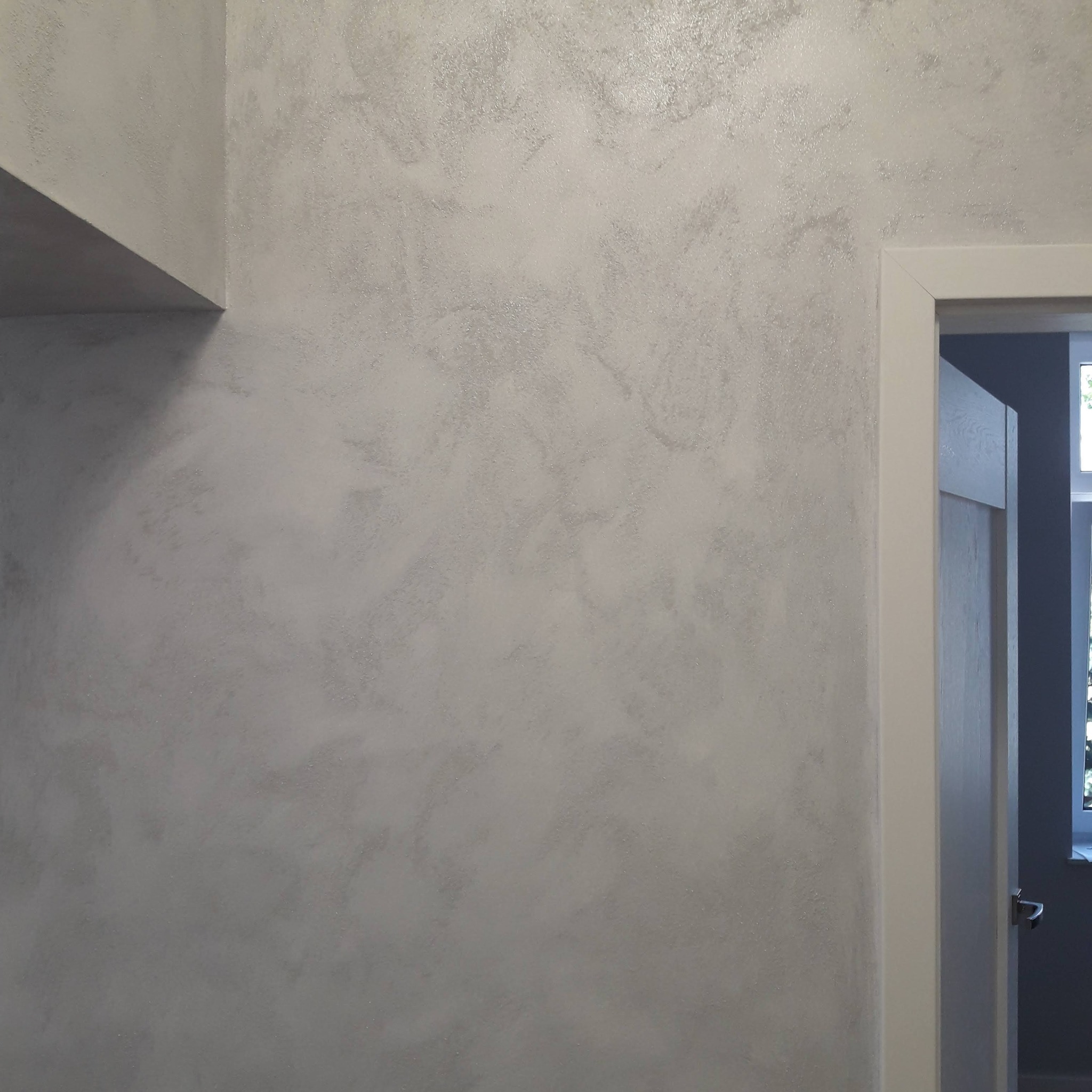 Ściana z dekoracyjnym, srebrzystym tynkiem strukturalnym, widoczny fragment sufitu i drzwi z białą ościeżnicą.