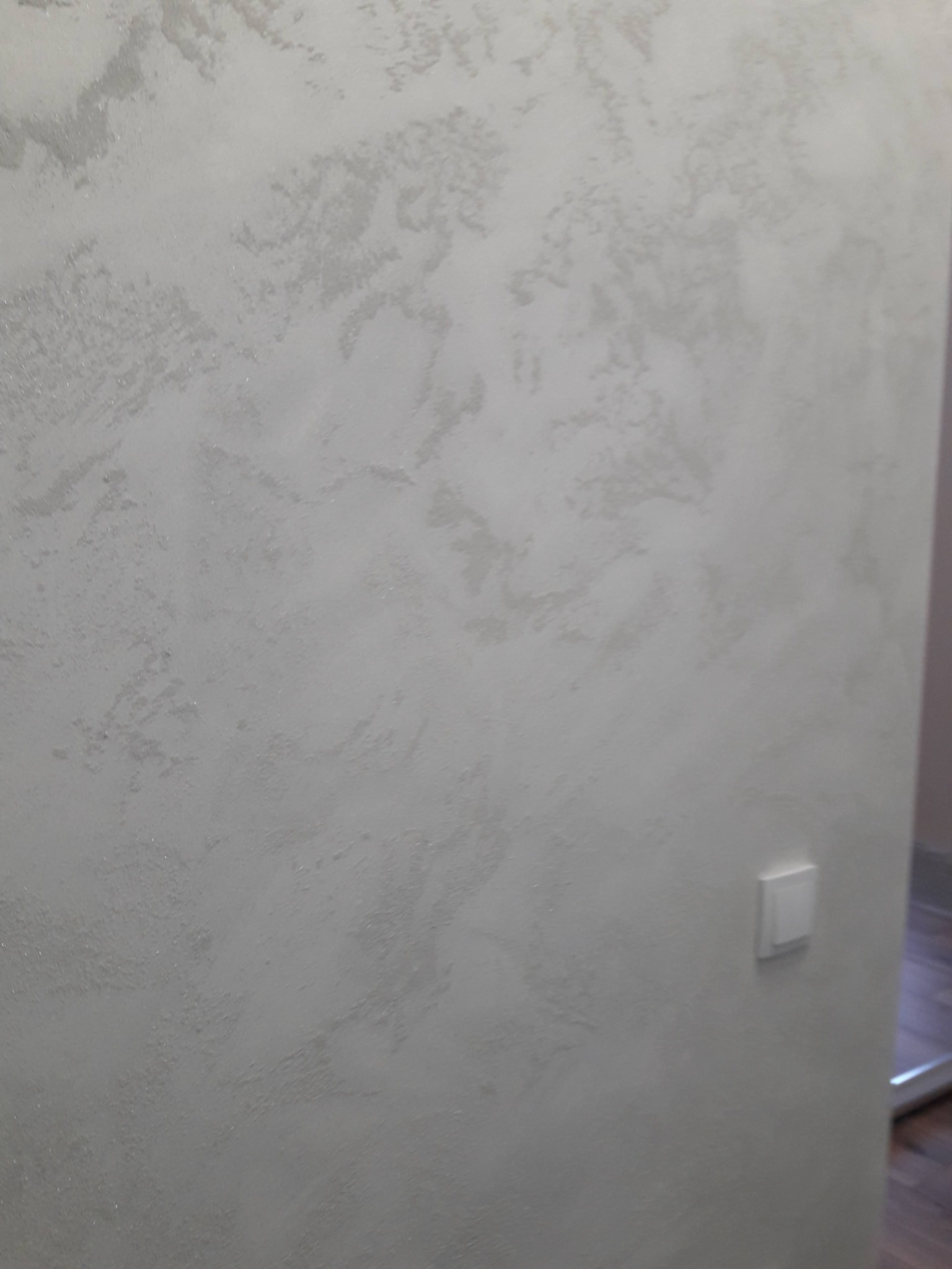 Ściana wewnętrzna z dekoracyjnym, srebrzystym tynkiem strukturalnym, obok włącznik światła