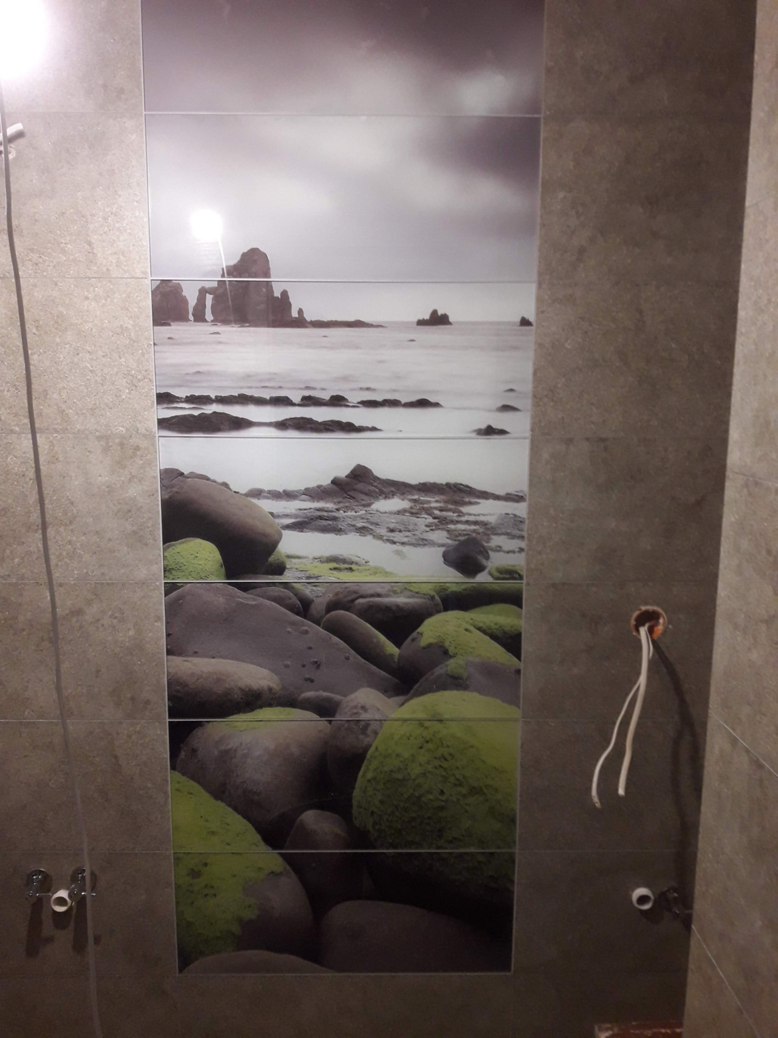 Wykończona ściana w łazience z szarymi płytkami, na których nadrukowano motyw morskiego krajobrazu ze skałami i mchem, widoczne wystające przewody elektryczne.