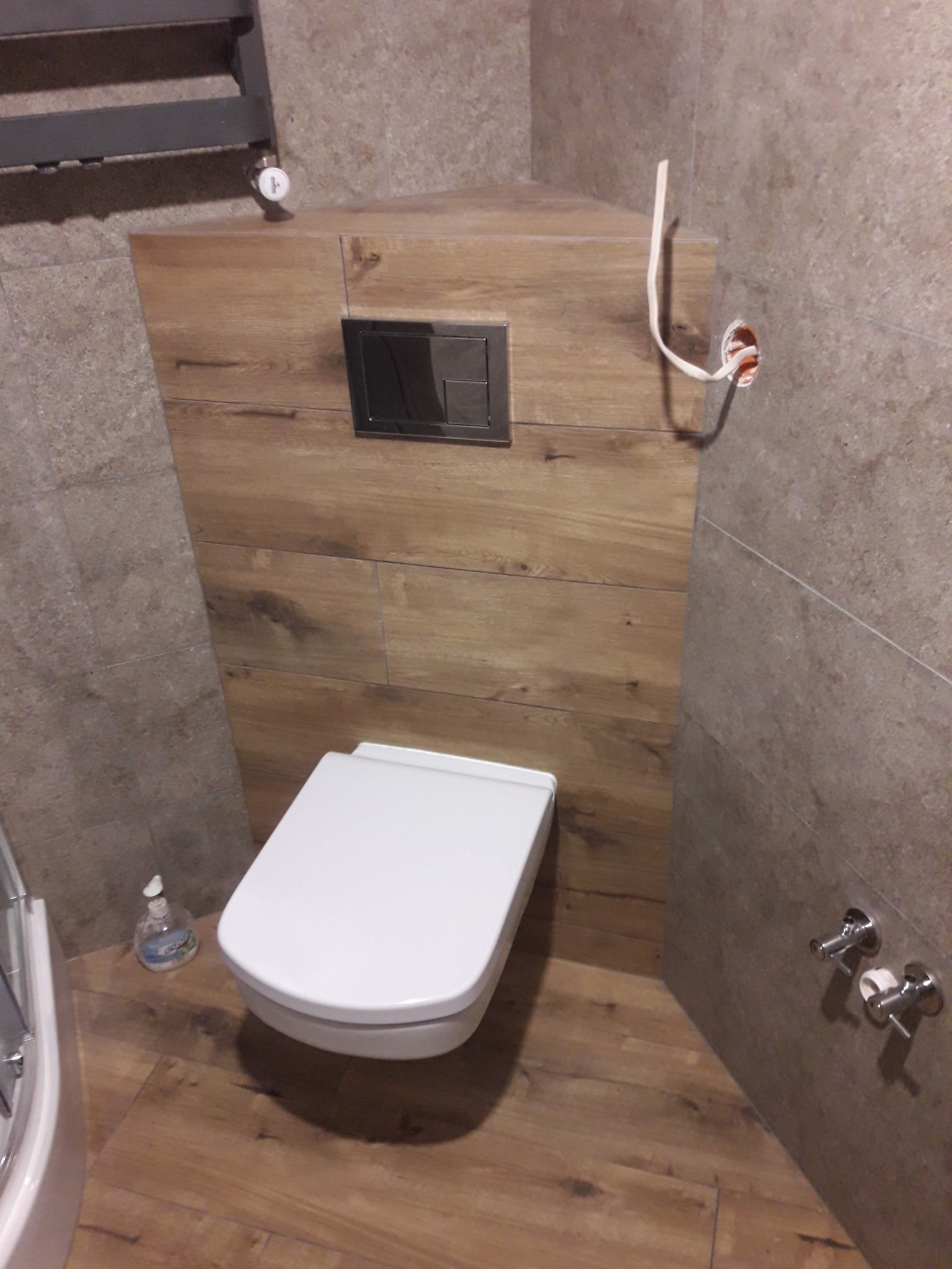 Nowoczesna, podwieszana toaleta w łazience wykończonej płytkami imitującymi drewno, z widocznymi przyłączami elektrycznymi i hydraulicznymi przed zabudową.